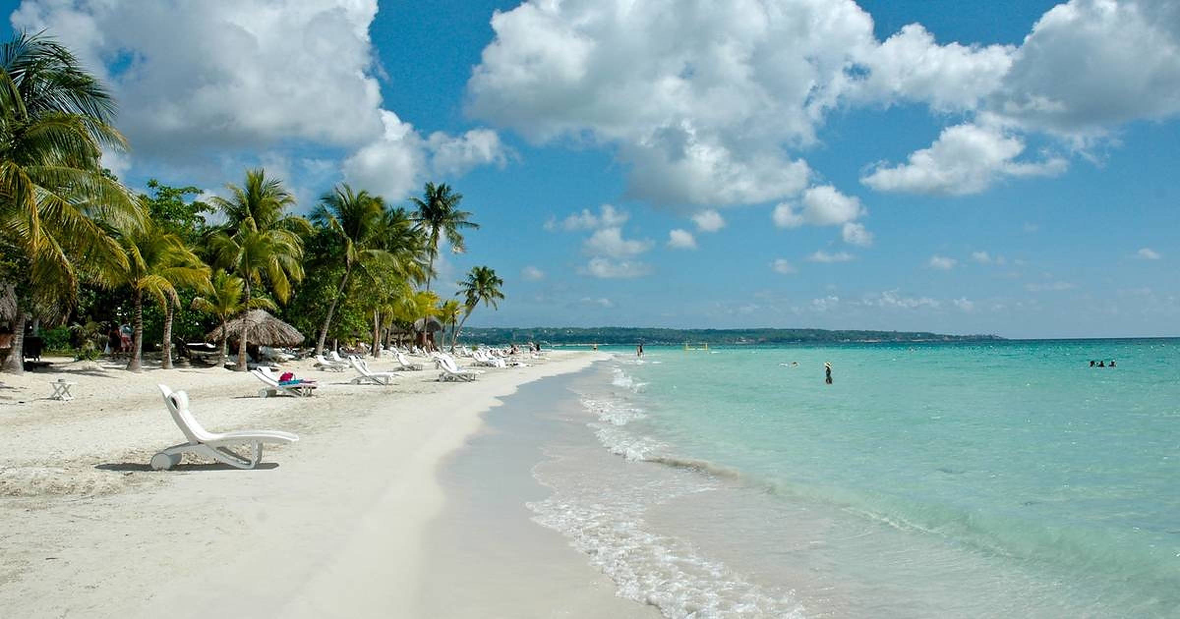 Jamaica Beach 桌布 壁紙 — 白色沙灘上排列著棕櫚樹和躺椅，在明亮的藍天下有蓬鬆的雲朵。