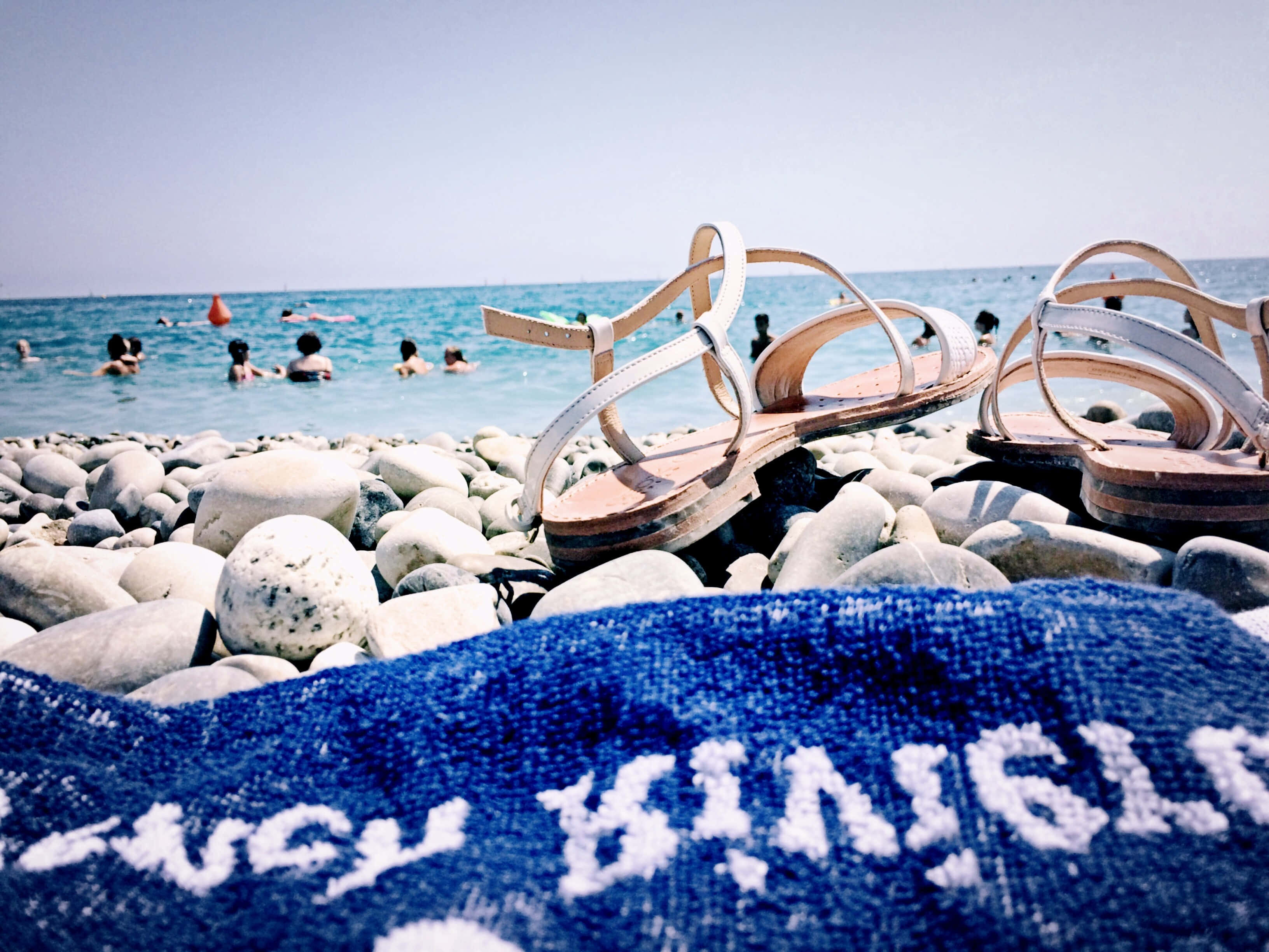 Beach Sandals 桌布 壁紙 — 白色涼鞋放在陽光照耀的鵝卵石上，旁邊有一條藍色毛巾，人們在晴朗天空下的碧綠海水中游泳。