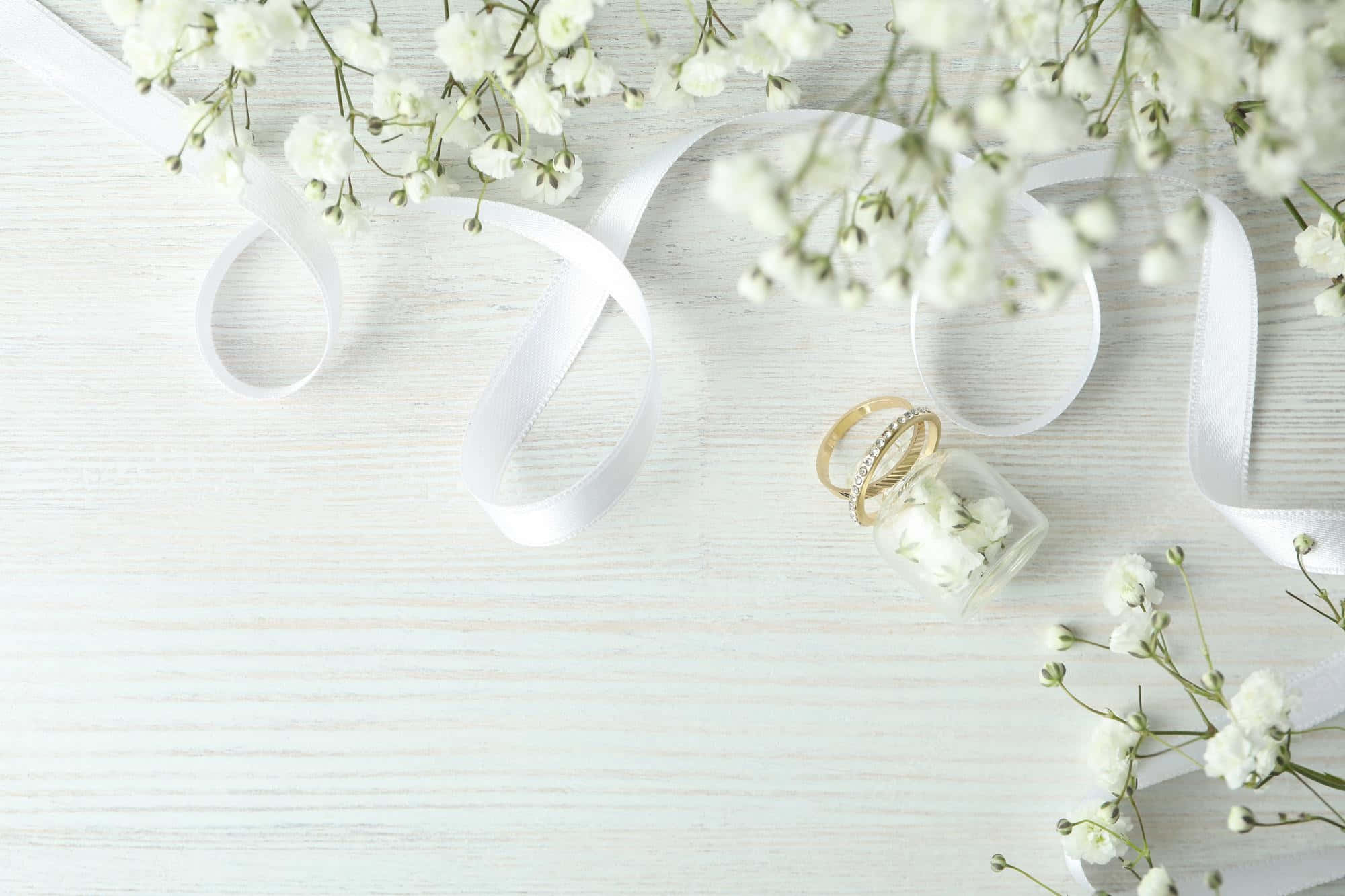 Wedding 桌布 壁紙 — 白色滿天星花朵和緞帶環繞著玻璃罐中的兩枚金色結婚戒指，置於白色木質表面上。