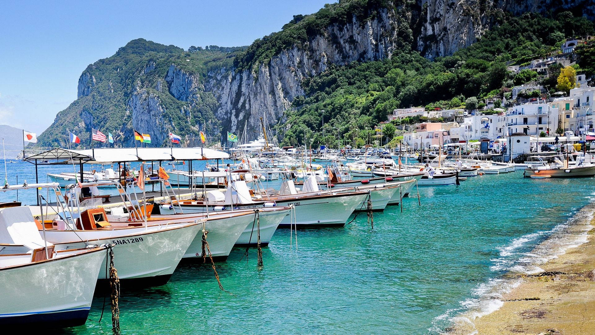 Capri 桌布 壁紙 — 白色漁船排列在碧綠色的海灣中，下方是陽光普照的懸崖村莊，色彩繽紛的旗幟隨風飄揚。