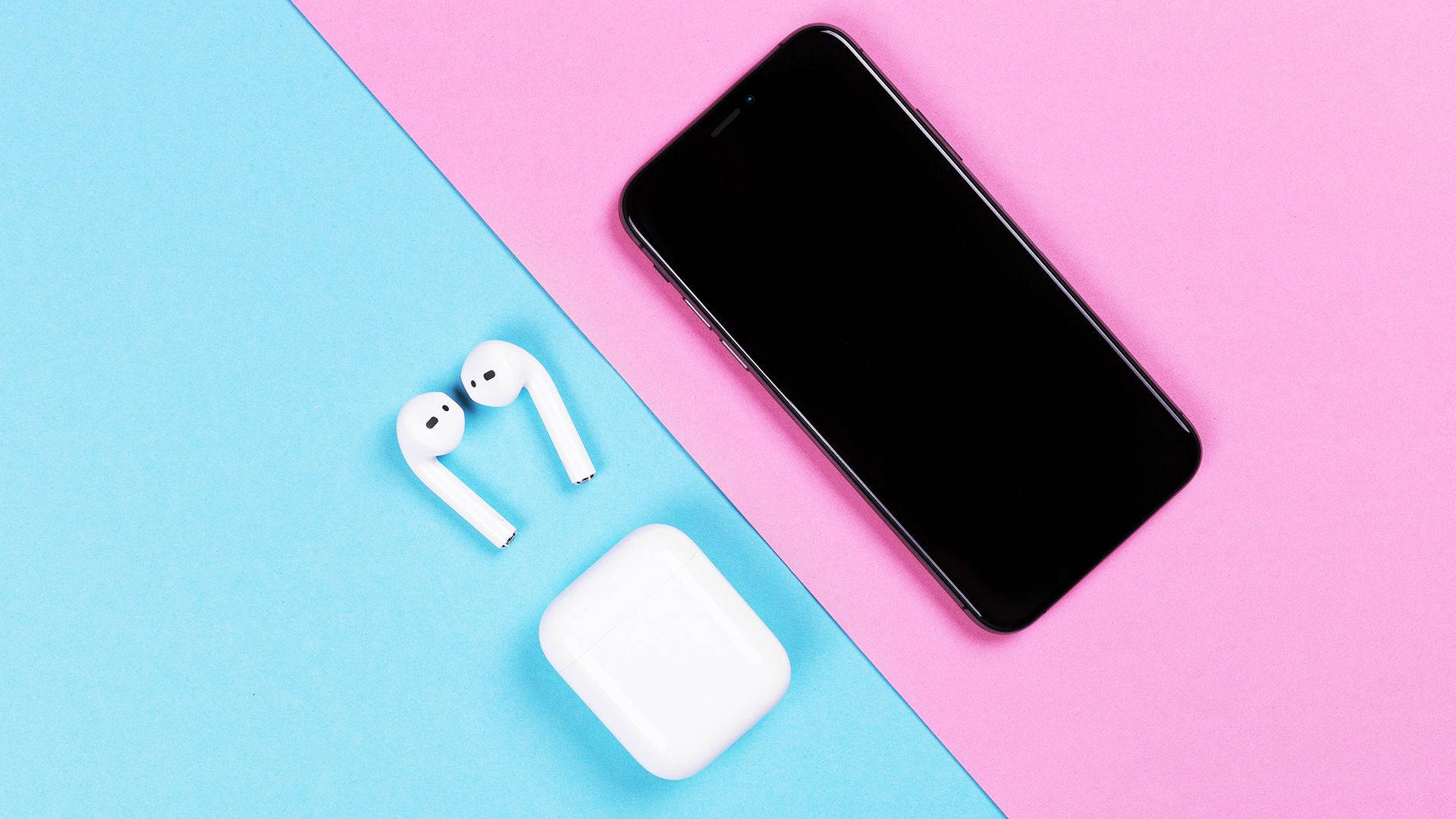 AirPods 桌布 壁紙 — 白色無線耳機和充電盒放在黑色智慧型手機旁邊，背景為粉紅色和藍色分割。
