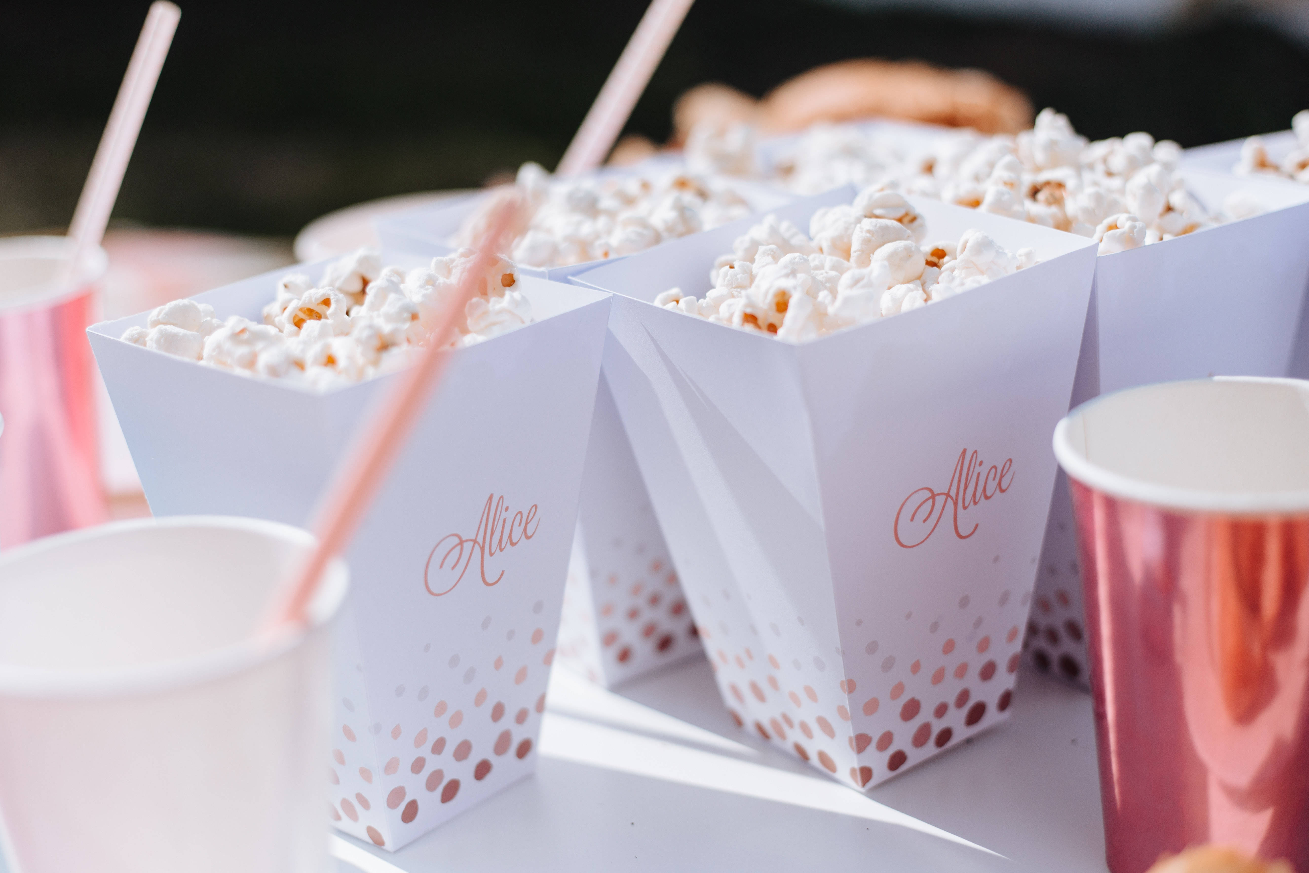Popcorn 桌布 壁紙 — 白色爆米花盒上有玫瑰金色圓點和 'Alice' 字樣，旁邊是閃亮的粉紅色杯子和吸管，放在明亮的桌子上。