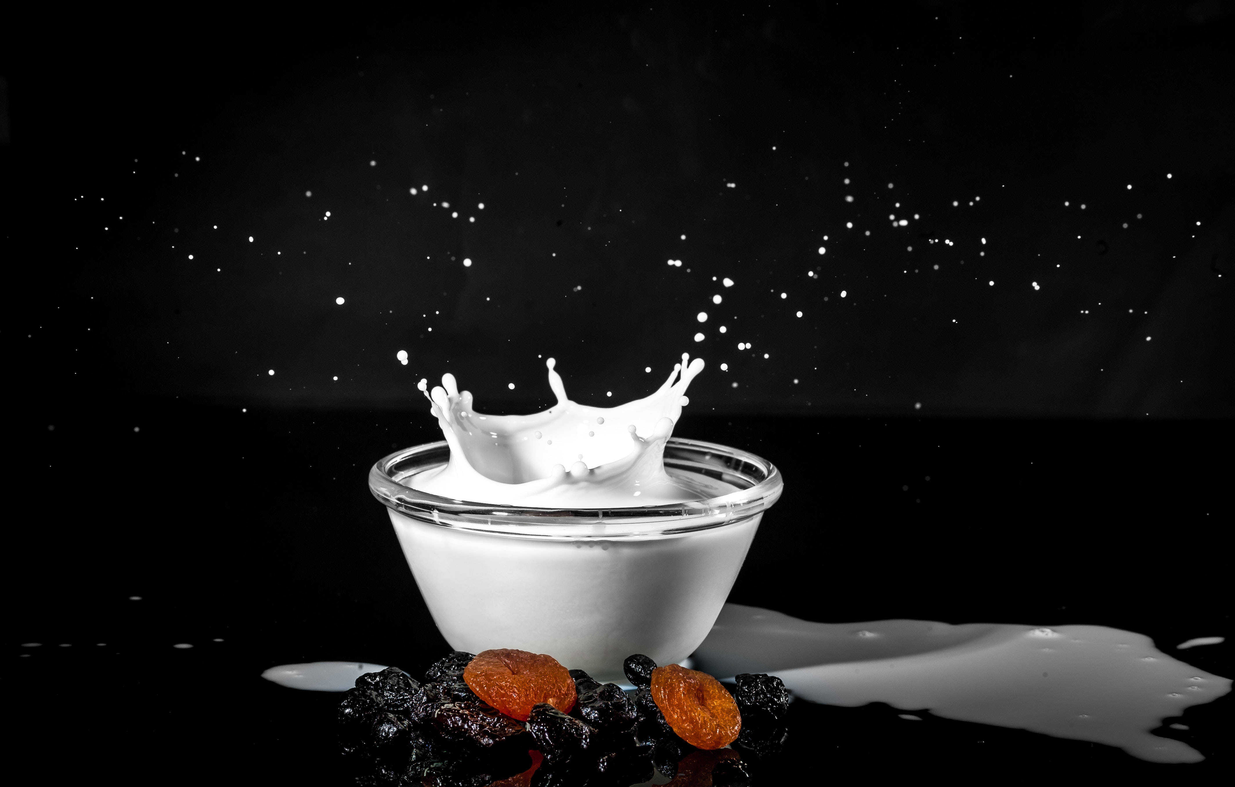 Milk 桌布 壁紙 — 白色牛奶從玻璃碗中動態噴濺而出，背景為純黑色，下方有散落的水滴和乾果。