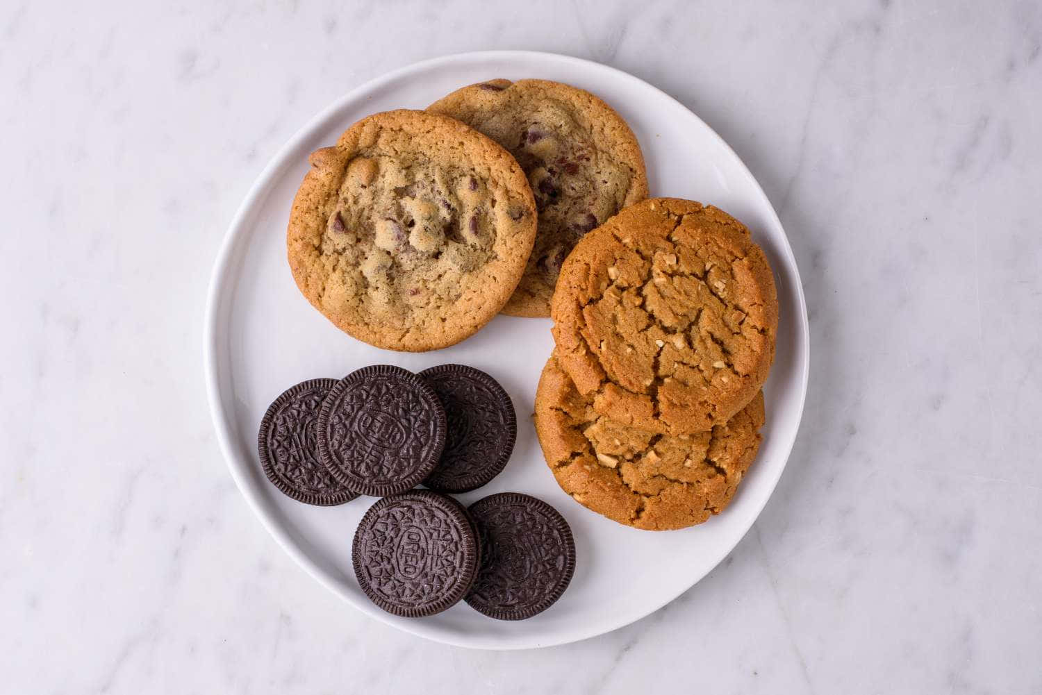 Cookie 桌布 壁紙 — 白色盤子上放著各種餅乾，包括巧克力曲奇、花生醬曲奇和Oreo曲奇，置於大理石表面上。