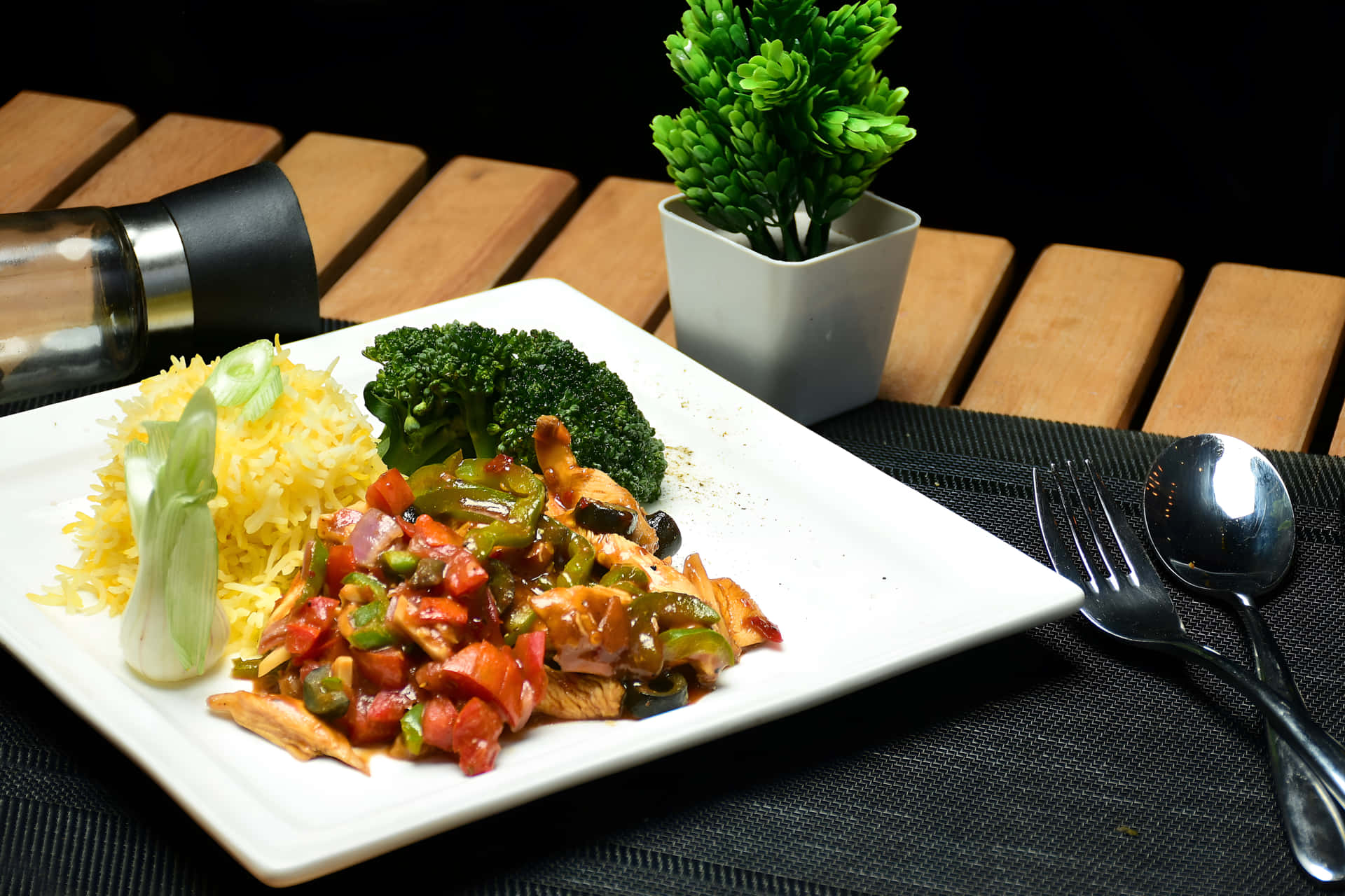 Chicken Chilli Dry with rice on a plate 壁紙 — 白色盤子上盛載色彩繽紛的炒雞肉、青花菜和黃色飯，放在木桌上，配有餐具和綠色植物。