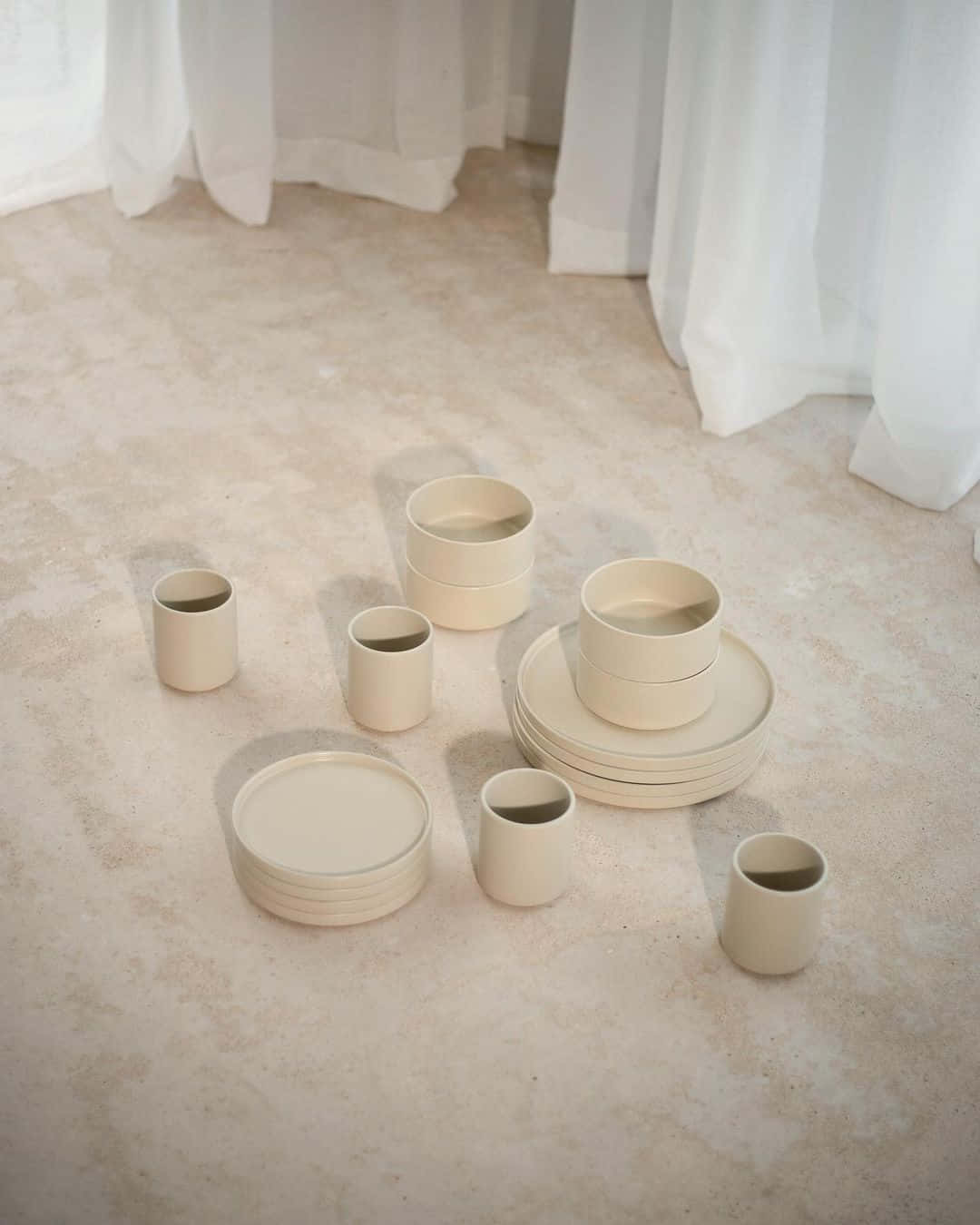 Ceramic 桌布 壁紙 — 白色窗簾附近柔和自然光下，質感地板上散落著極簡米色陶瓷餐具。
