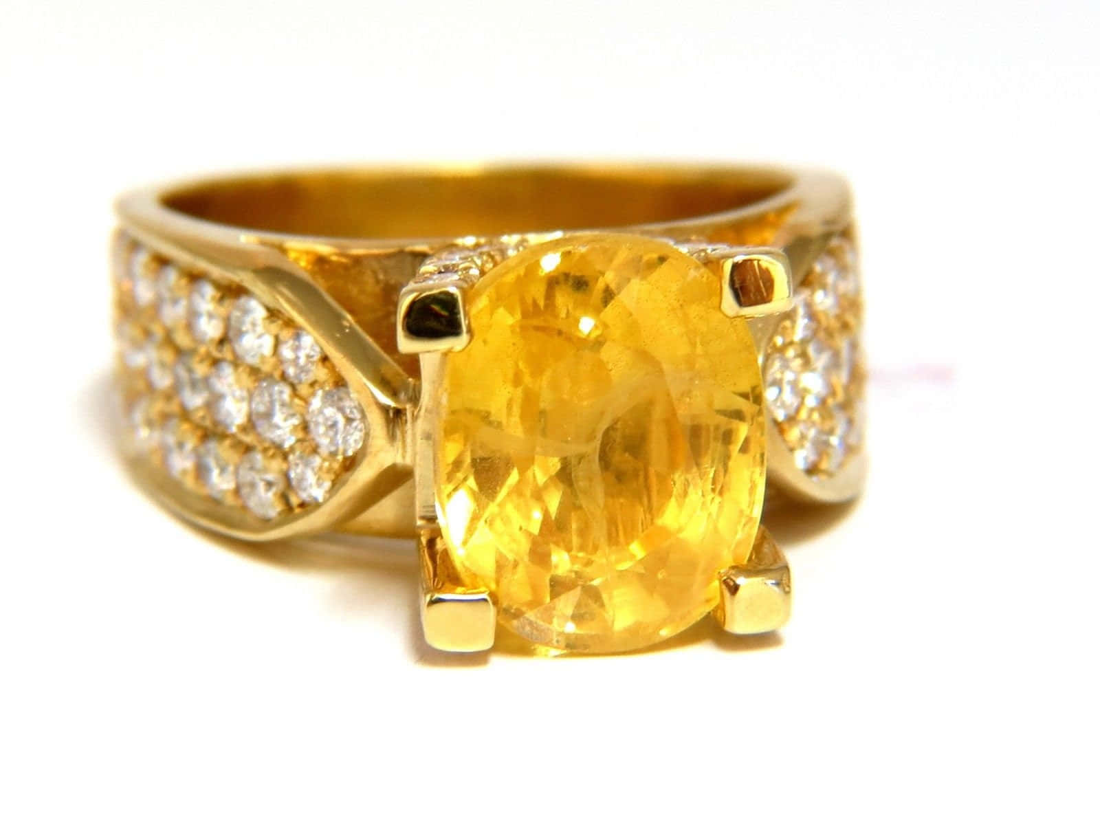 Yellow Sapphire 桌布 壁紙 — 白色背景上，一枚黃金戒指的特寫，戒指上有一顆大型橢圓形黃色寶石，周圍鑲嵌著較小的鑽石。