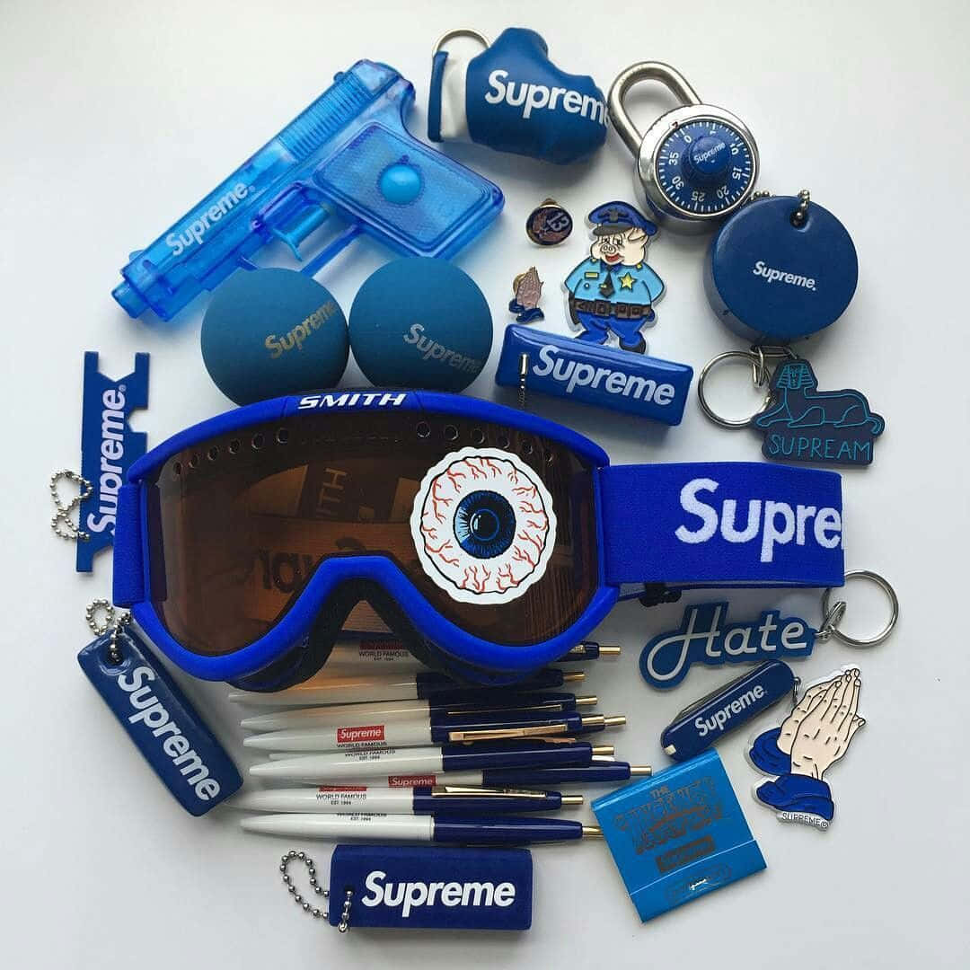 blue supreme 桌布 壁紙 — 白色背景上藍色Supreme品牌商品的平鋪擺放，包括帶有眼睛設計的護目鏡、筆、鑰匙圈和玩具槍。
