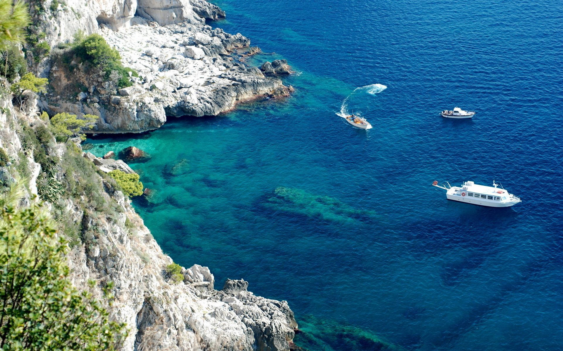 Capri 桌布 壁紙 — 白色船隻在岩石懸崖和茂密綠意之間滑行，俯瞰碧綠海灣的航拍圖。