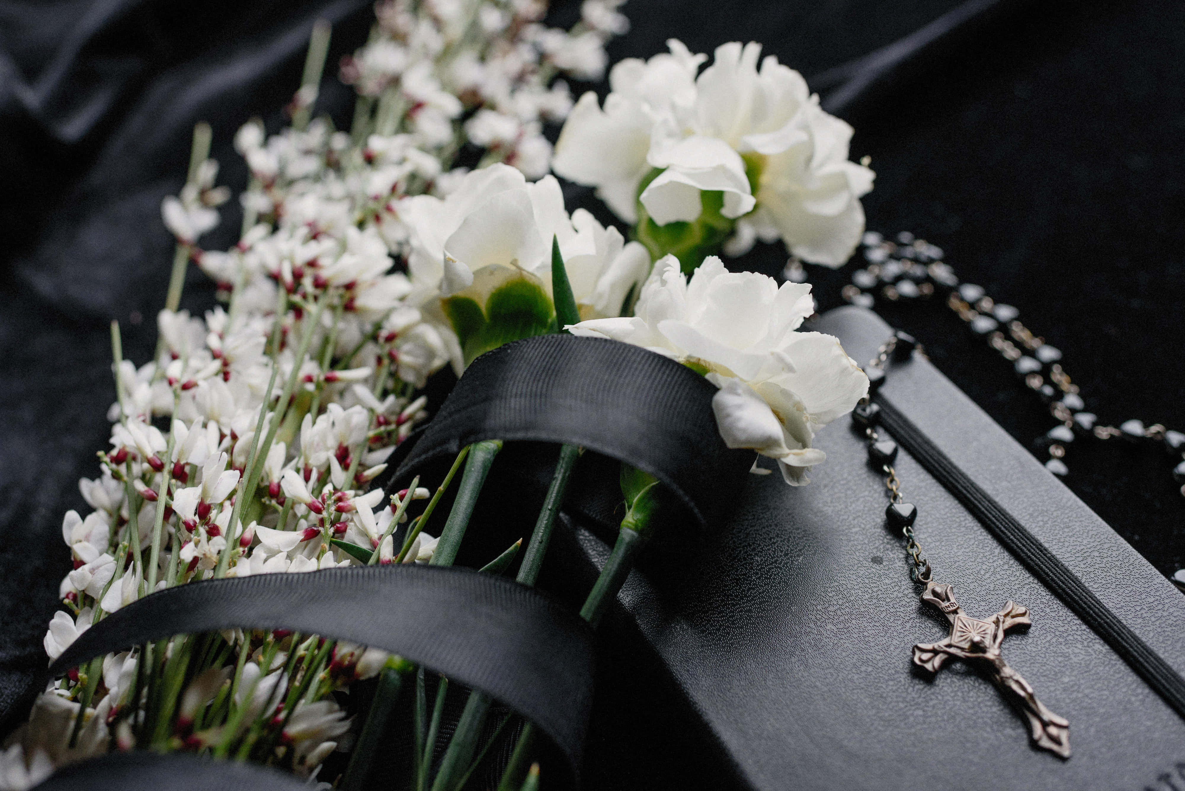 Memorial 桌布 壁紙 — 白色花朵和玫瑰經唸珠莊嚴地放在一本黑色書本上，背景是深色布料。