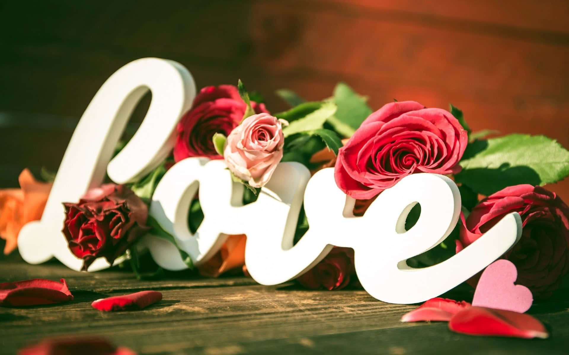 Valentines 桌布 壁紙 — 白色草書 'love' 字母依偎在充滿活力的紅粉玫瑰之間，置於質樸的木質表面上。