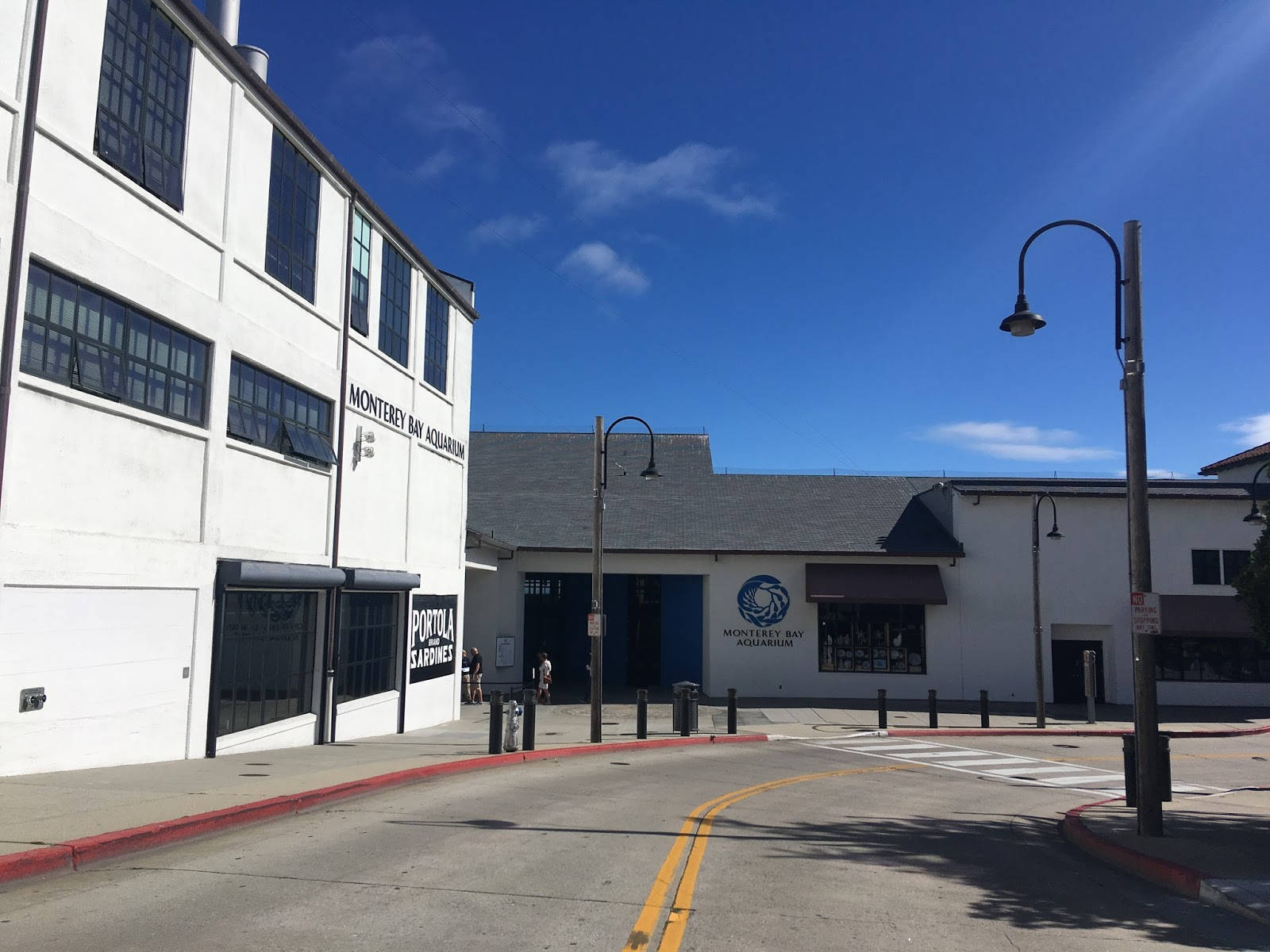 Road 桌布 壁紙 — 白色蒙特雷灣水族館建築，在明亮的藍天下有散落的雲朵和投射陰影的路燈。