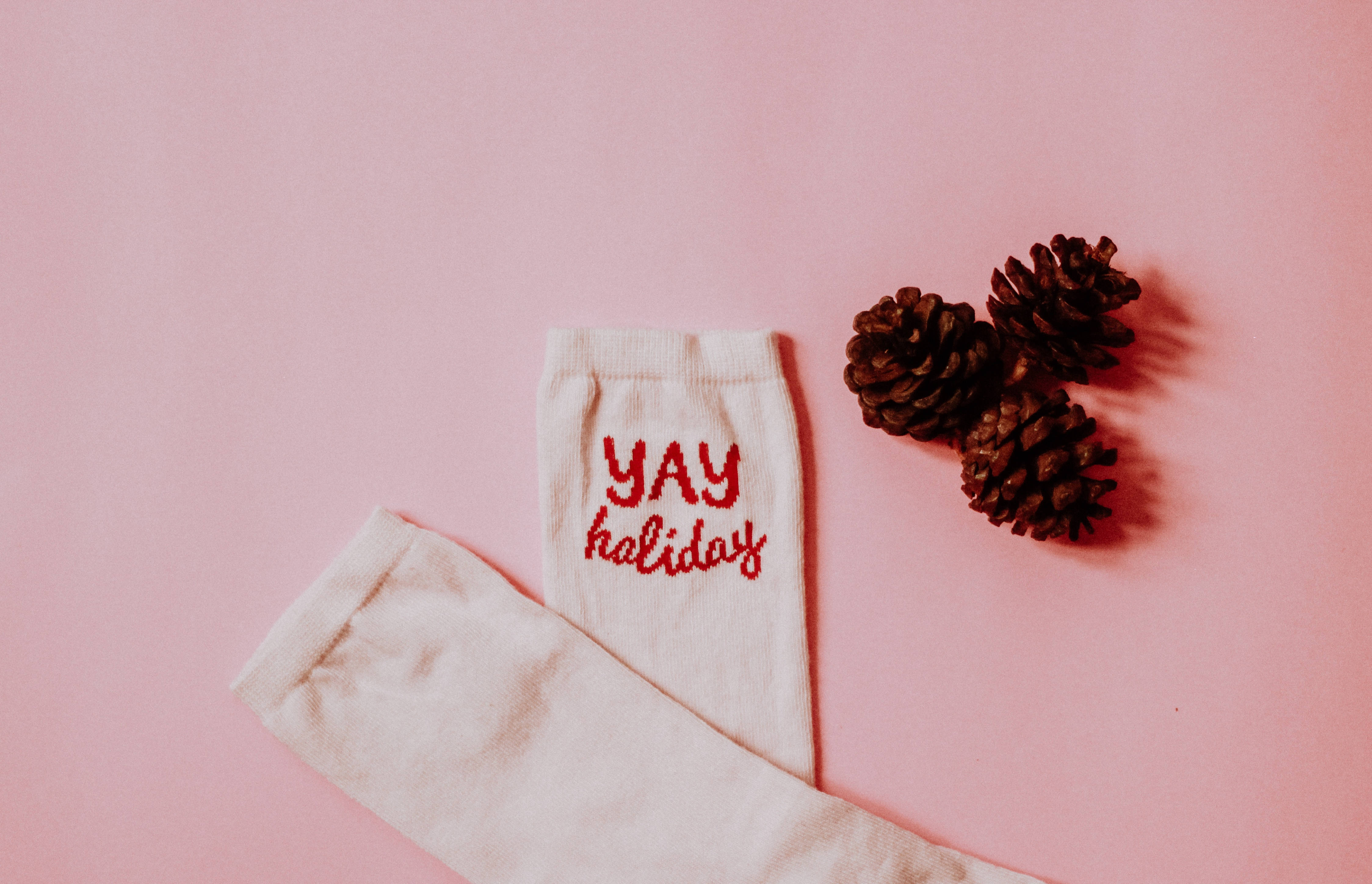 Christmas Vacation 桌布 壁紙 — 白色襪子上有紅色 'YAY holiday' 文字及三個松果，置於柔軟粉紅背景上。