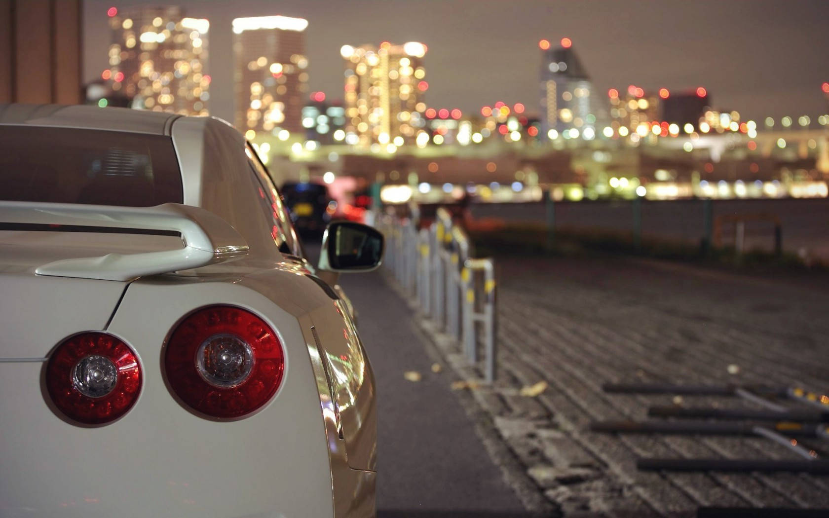 桌布 壁紙 — 白色跑車的車尾在都市燈光下閃耀，背景是模糊的發光摩天大樓天際線。