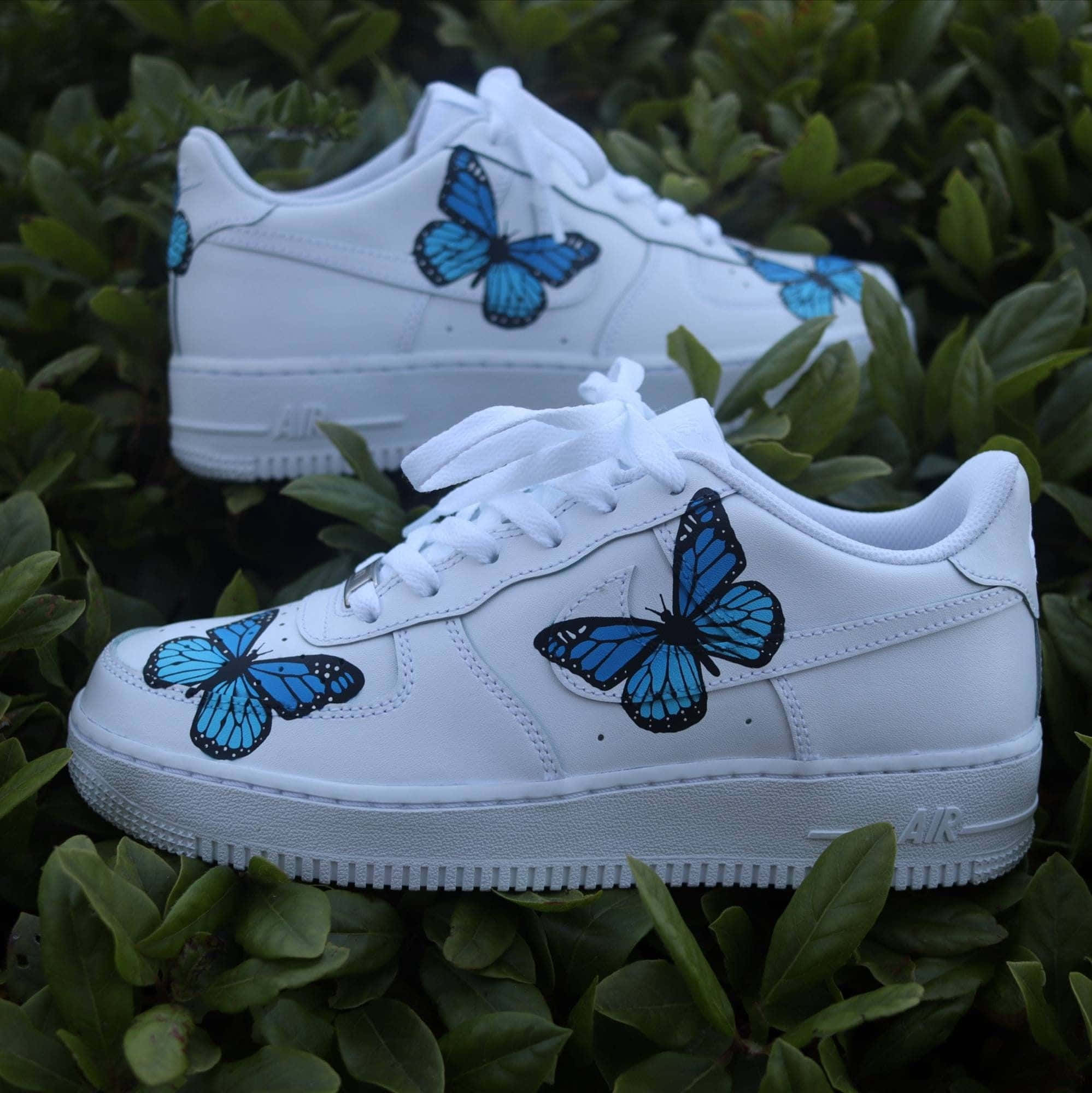 Nike Air Force 桌布 壁紙 — 白色運動鞋點綴著鮮豔的藍色蝴蝶，放置在茂密的綠色樹葉中。