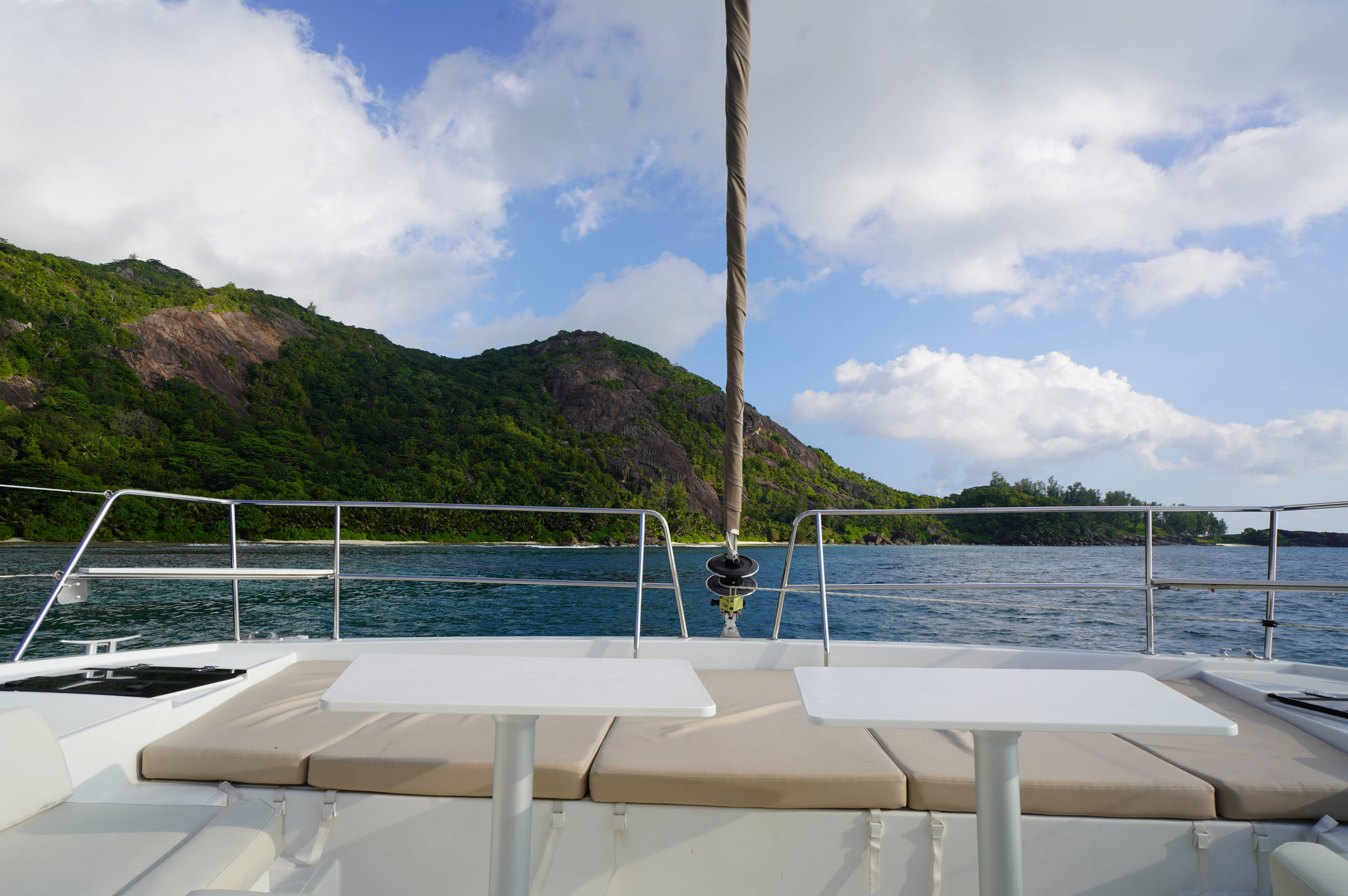 Seychelles 桌布 壁紙 — 白色雙體船甲板配米色軟墊，俯瞰在部分多雲的藍天下呈現碧綠色的海水和鬱鬱蔥蔥的綠色山丘。