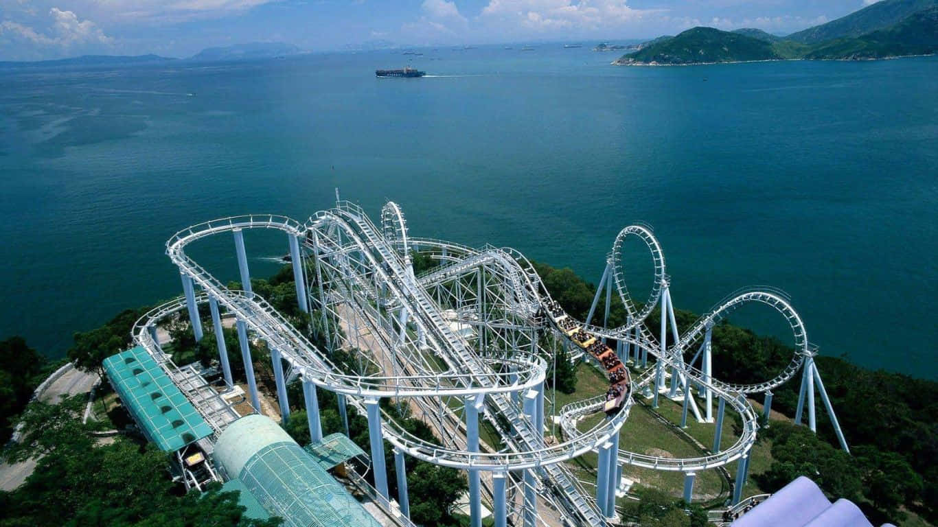 Roller Coaster 桌布 壁紙 — 白色雲霄飛車在鬱鬱蔥蔥的綠色山丘上蜿蜒曲折，旁邊是碧綠的海洋和遠處島嶼的鳥瞰圖。