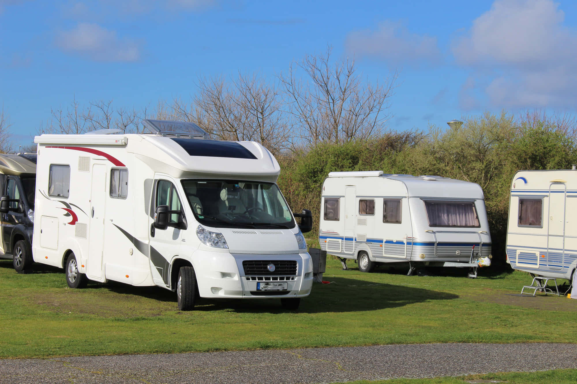 Caravan 桌布 壁紙 — 白色露營車停泊在草地上，背景是明亮的藍天、散落的雲朵和無葉的樹木。