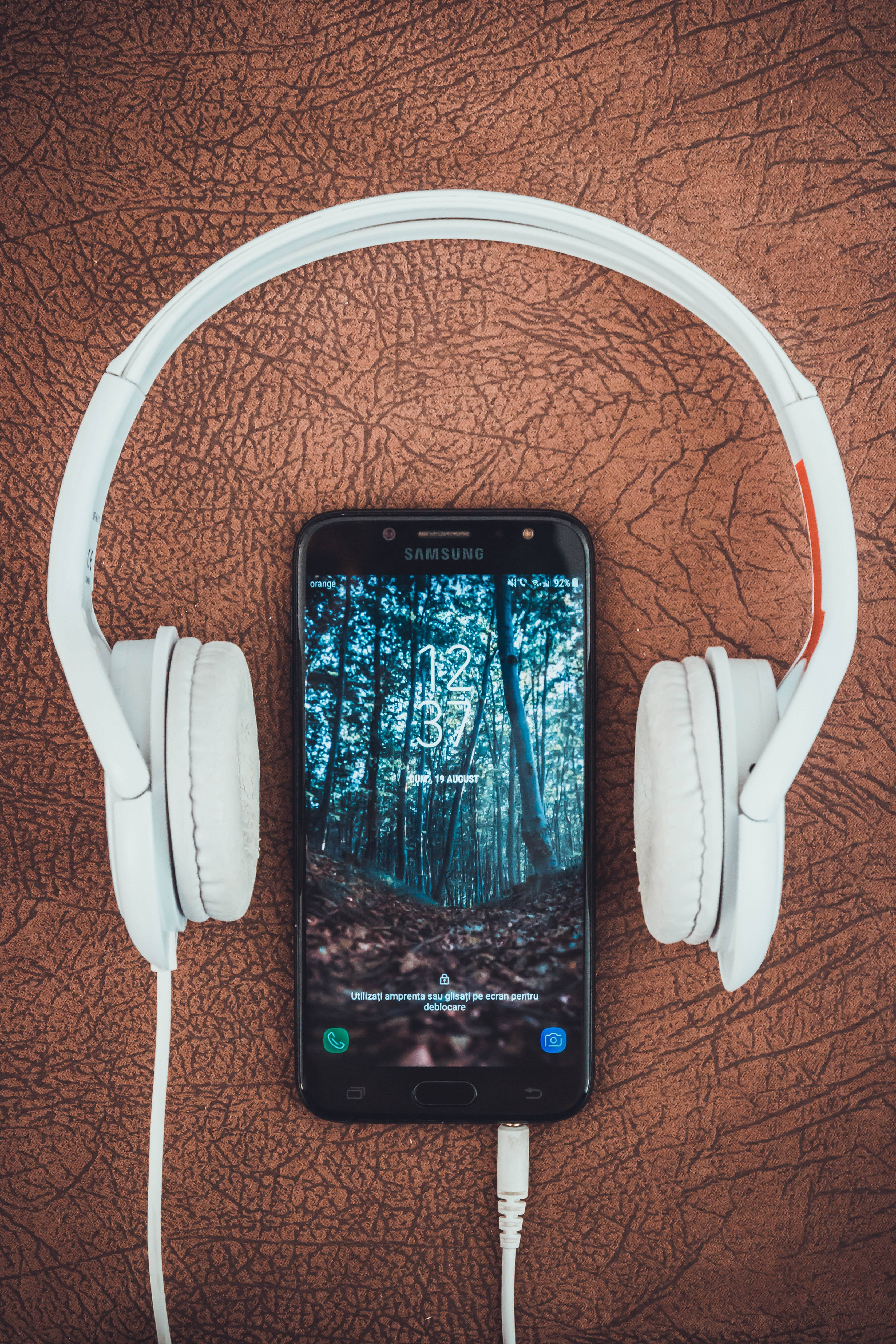 音樂 桌布 壁紙 — 白色頭戴式耳機放在一支顯示森林桌布的智慧型手機上，手機置於有紋理的棕色表面。