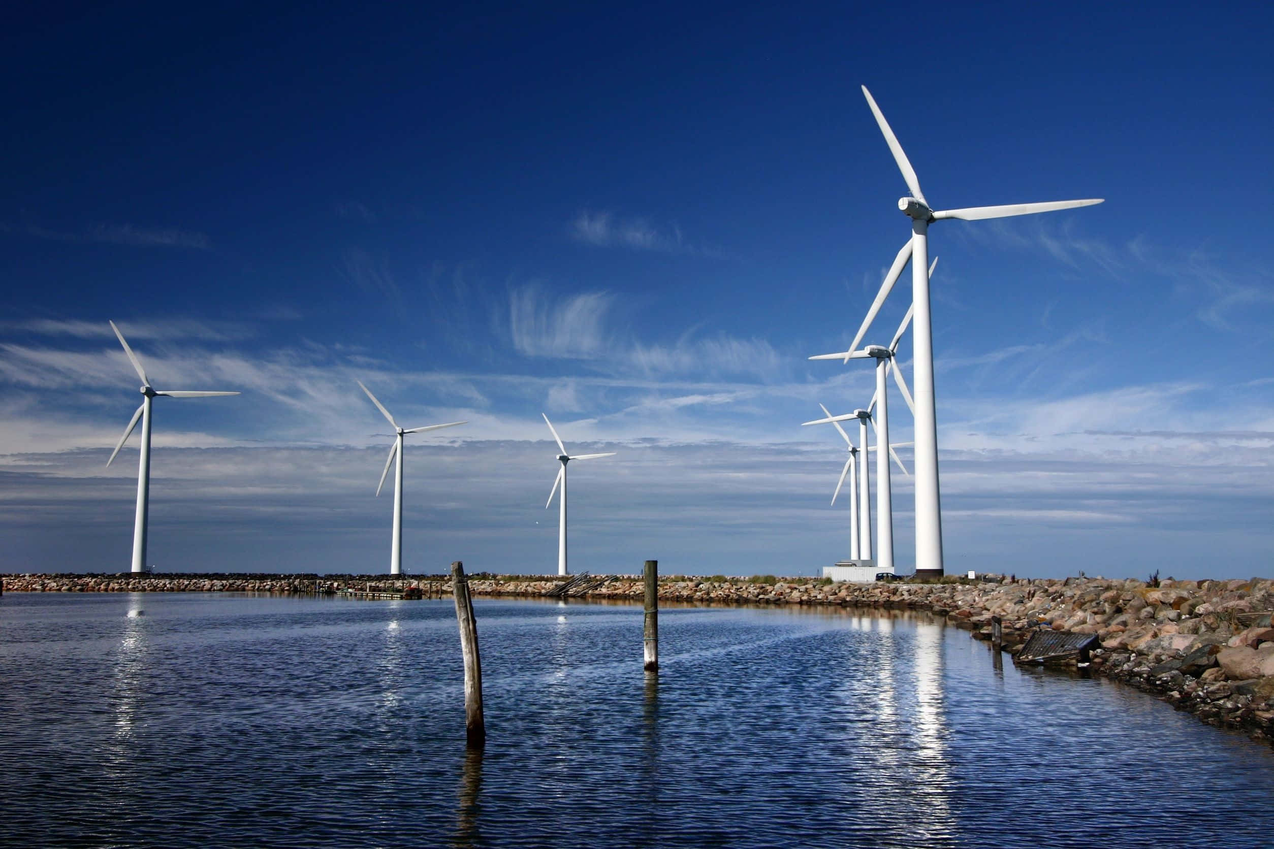 Energy 桌布 壁紙 — 白色風力發電機在清澈的藍天下高高聳立，倒映在岩石海岸線附近的平靜水面上。