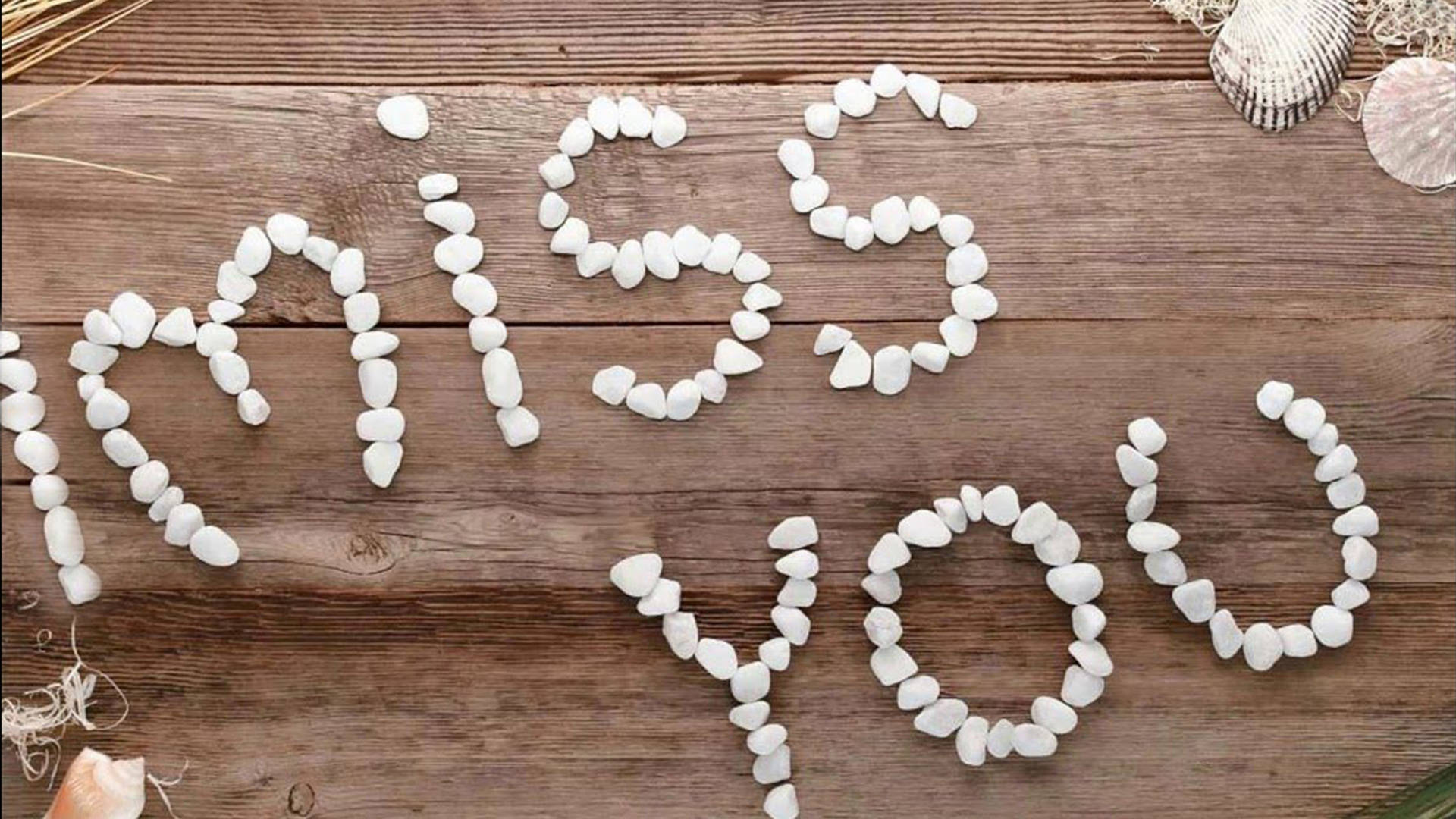 我想你 桌布 壁紙 — 白色鵝卵石在風化的木質表面上拼出「miss you」，旁邊有貝殼和乾草。