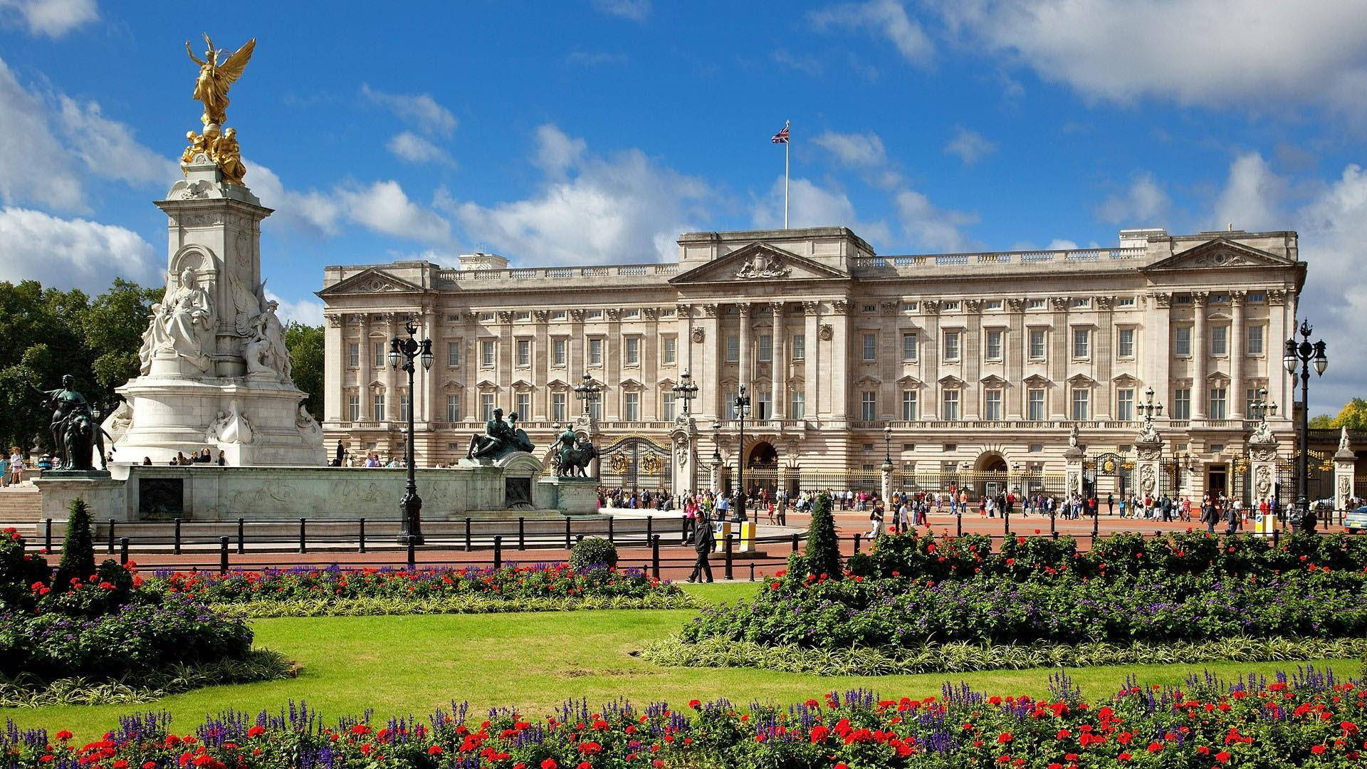 England 桌布 壁紙 — 白金漢宮在晴朗藍天和蓬鬆白雲下巍然聳立，周圍環繞著色彩繽紛的花壇和維多利亞紀念碑。