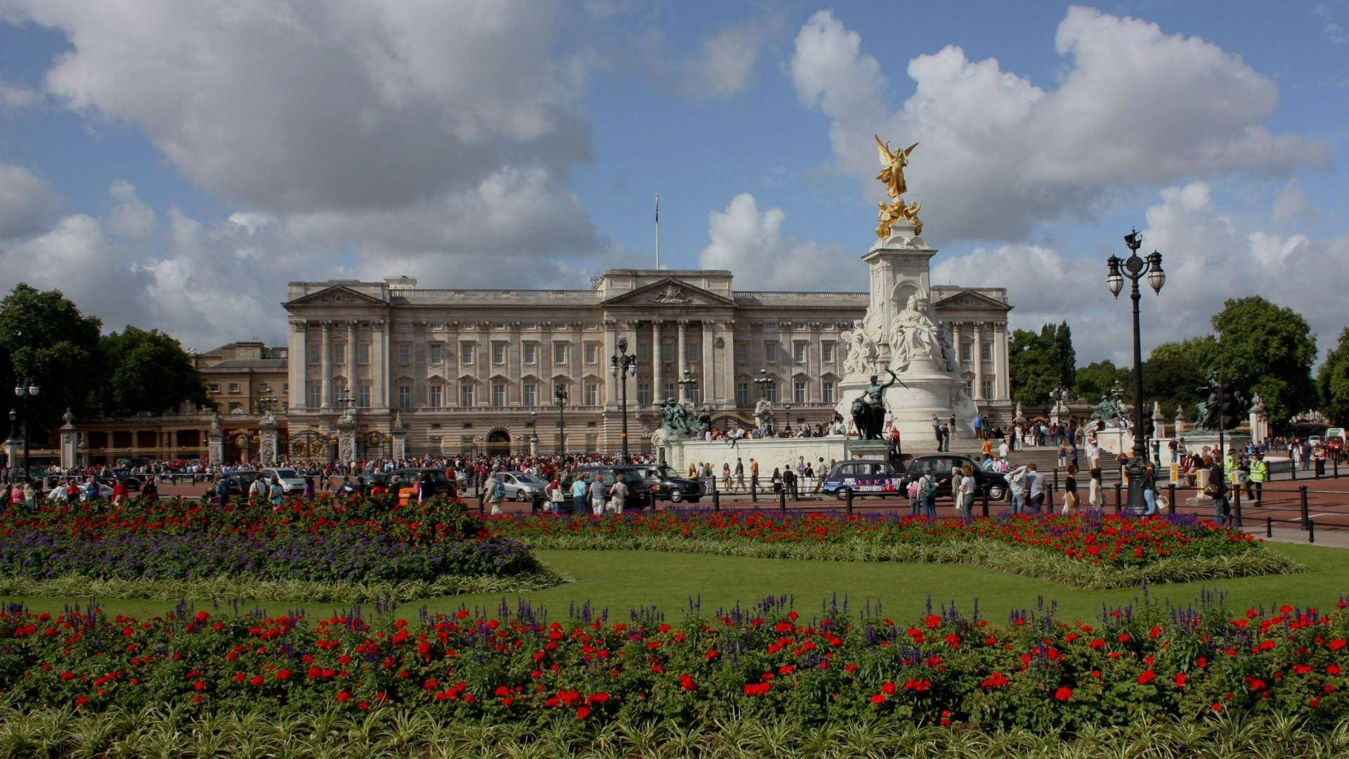 England 桌布 壁紙 — 白金漢宮在部分多雲的天空下雄偉矗立，周圍有鮮豔的花壇和聚集在維多利亞紀念碑附近的人群。