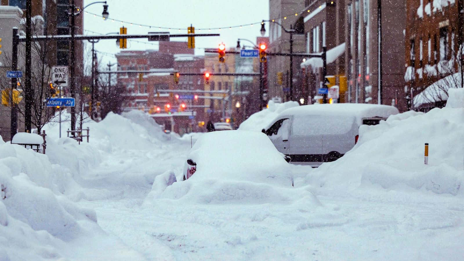 blizzard 桌布 壁紙 — 白雪覆蓋城市街道，汽車被埋在雪堆下，交通燈在冬季風暴中發出紅光。