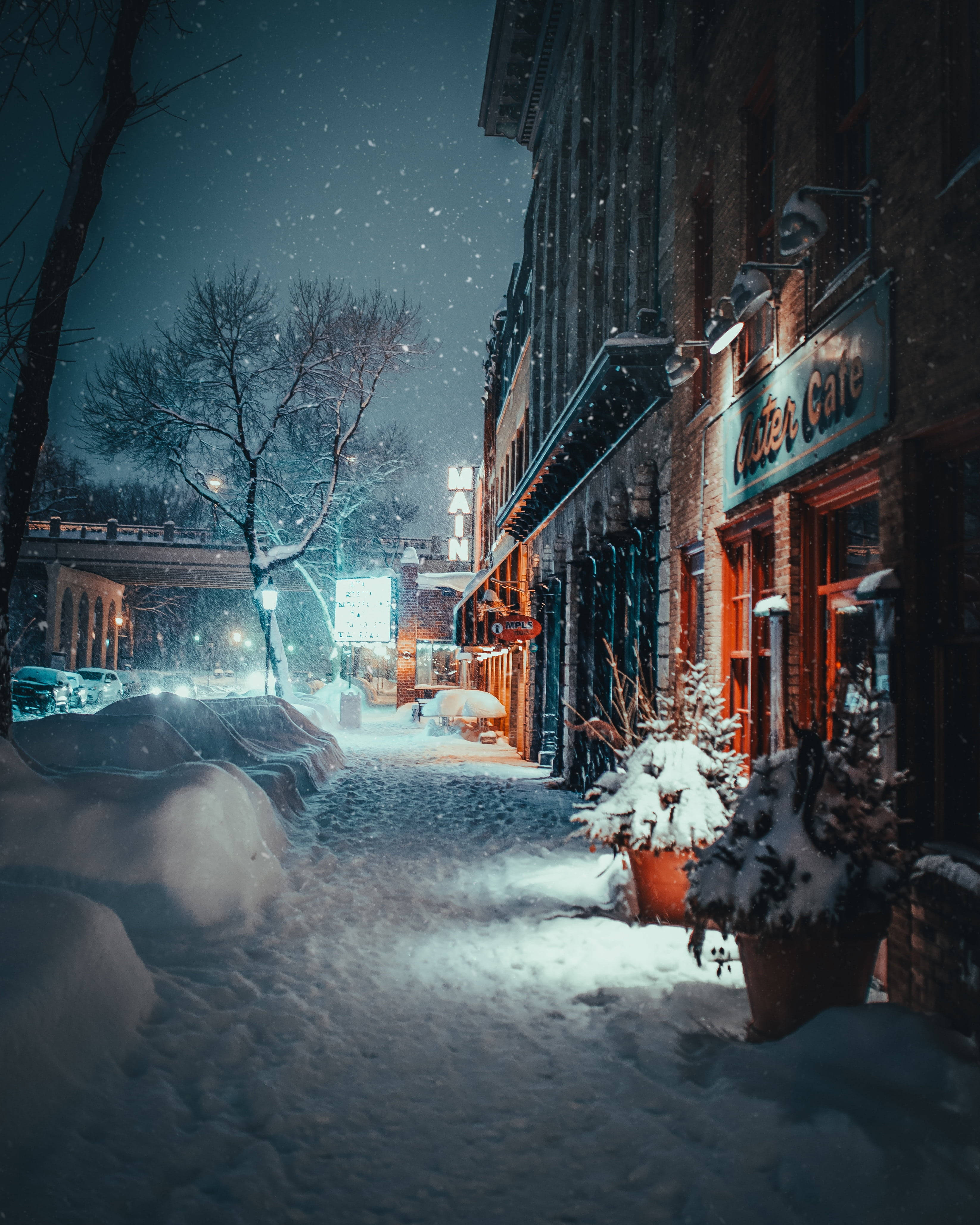 冬天 桌布 壁紙 — 白雪覆蓋寧靜的城市街道，夜晚時分，店舖櫥窗的溫暖燈光在飄落的雪花和漆黑的天空中閃耀。