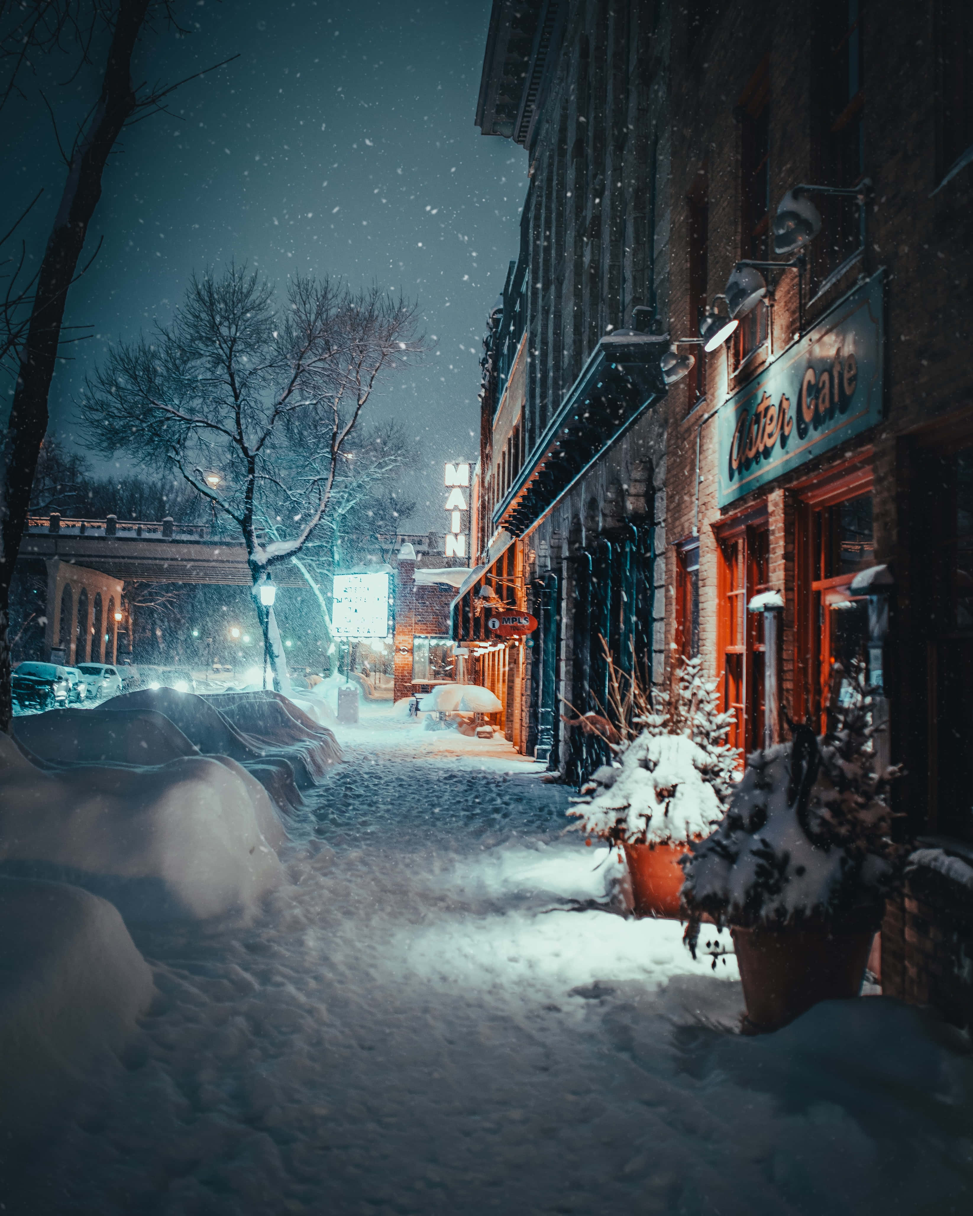 Christmas Scene 桌布 壁紙 — 白雪覆蓋寧靜的城市街道，夜晚溫暖的燈光從商店櫥窗中閃爍，飄落的雪花照亮了場景。