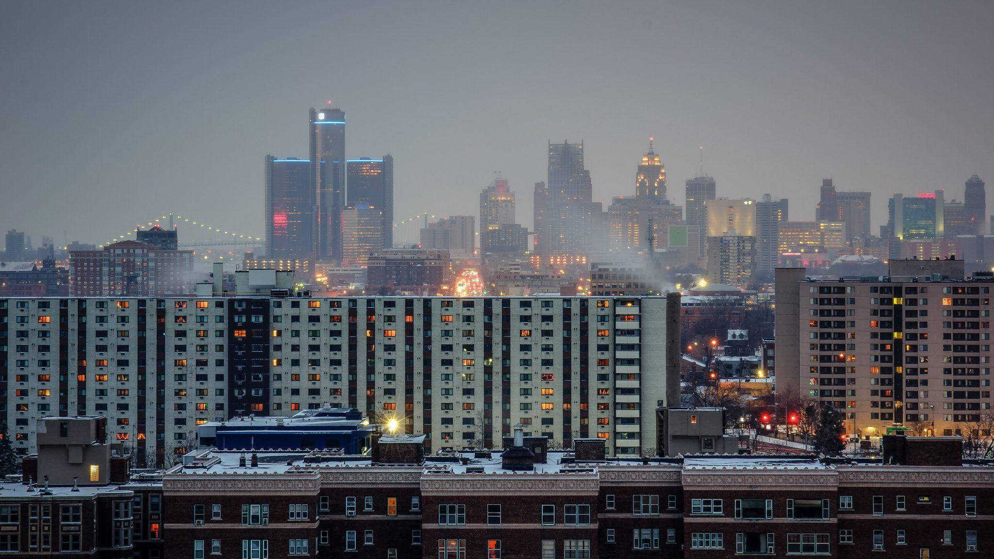 detroit 桌布 壁紙 — 白雪覆蓋屋頂，城市燈光在朦朧的暮色天空中溫暖地閃耀，映襯著密集的城市天際線。