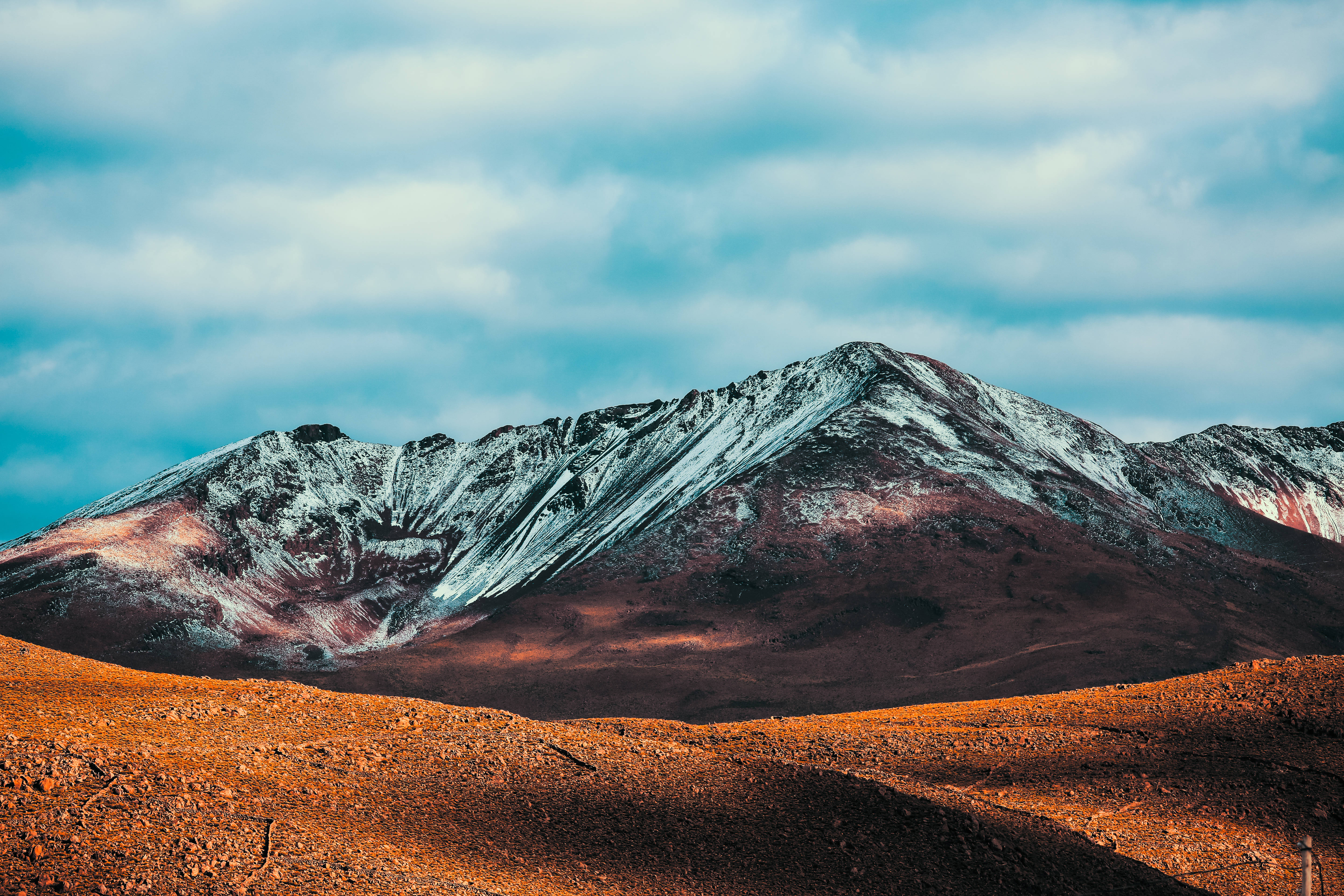 Bolivia 桌布 壁紙 — 白雪覆蓋崎嶇的山峰，多雲的天空下是乾燥的橙紅色地形。