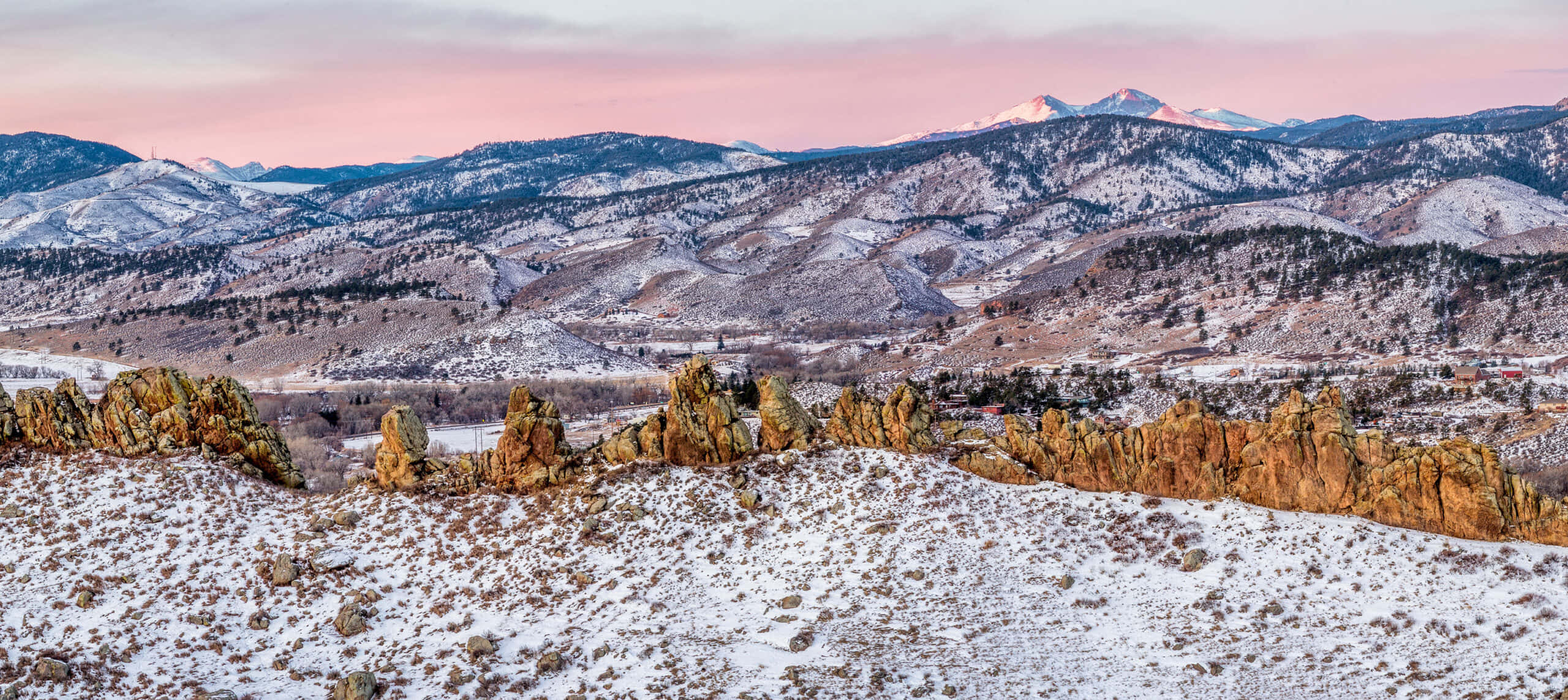 Colorado Mountain 桌布 壁紙 — 白雪覆蓋崎嶇的岩石地貌和起伏的丘陵，在柔和的粉紅色和橙色暮色天空下，遠處有積雪的山峰。