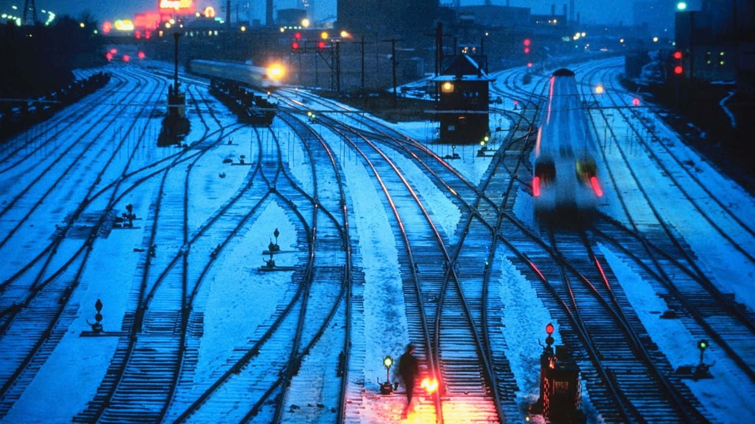 1536x864 桌布 壁紙 — 白雪覆蓋廣闊的鐵路調車場，夜晚火車在信號燈的紅黃光芒中滑行。