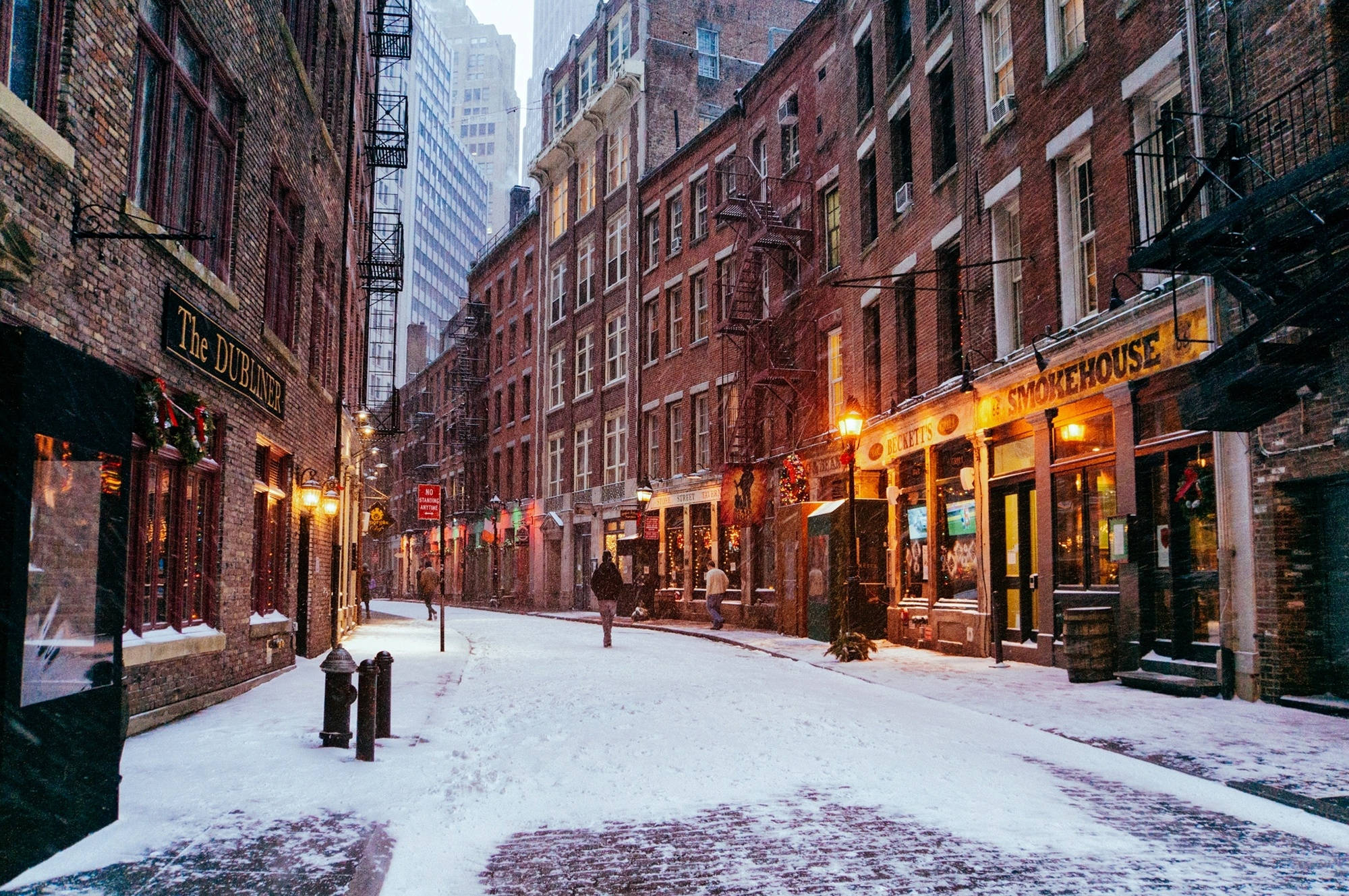 雪 桌布 壁紙 — 白雪覆蓋歷史建築之間狹窄的磚砌小巷，溫暖發光的商店門面和逃生樓梯。