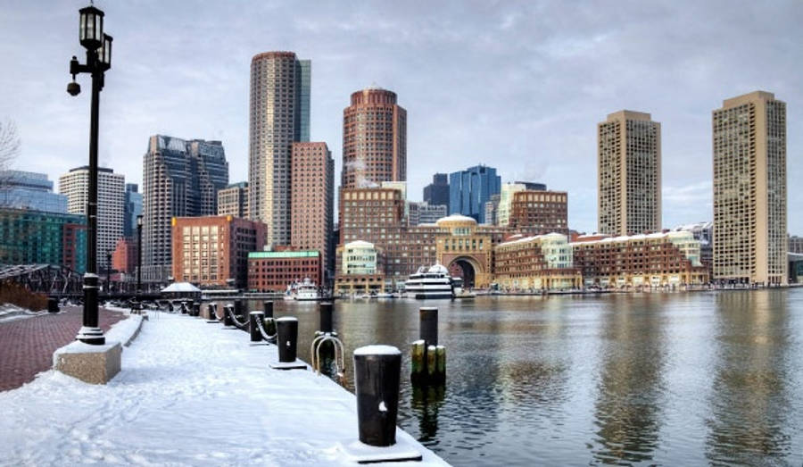 Massachusetts 桌布 壁紙 — 白雪覆蓋海濱長廊，現代摩天大樓在陰天之下倒映在平靜的港灣水面上。