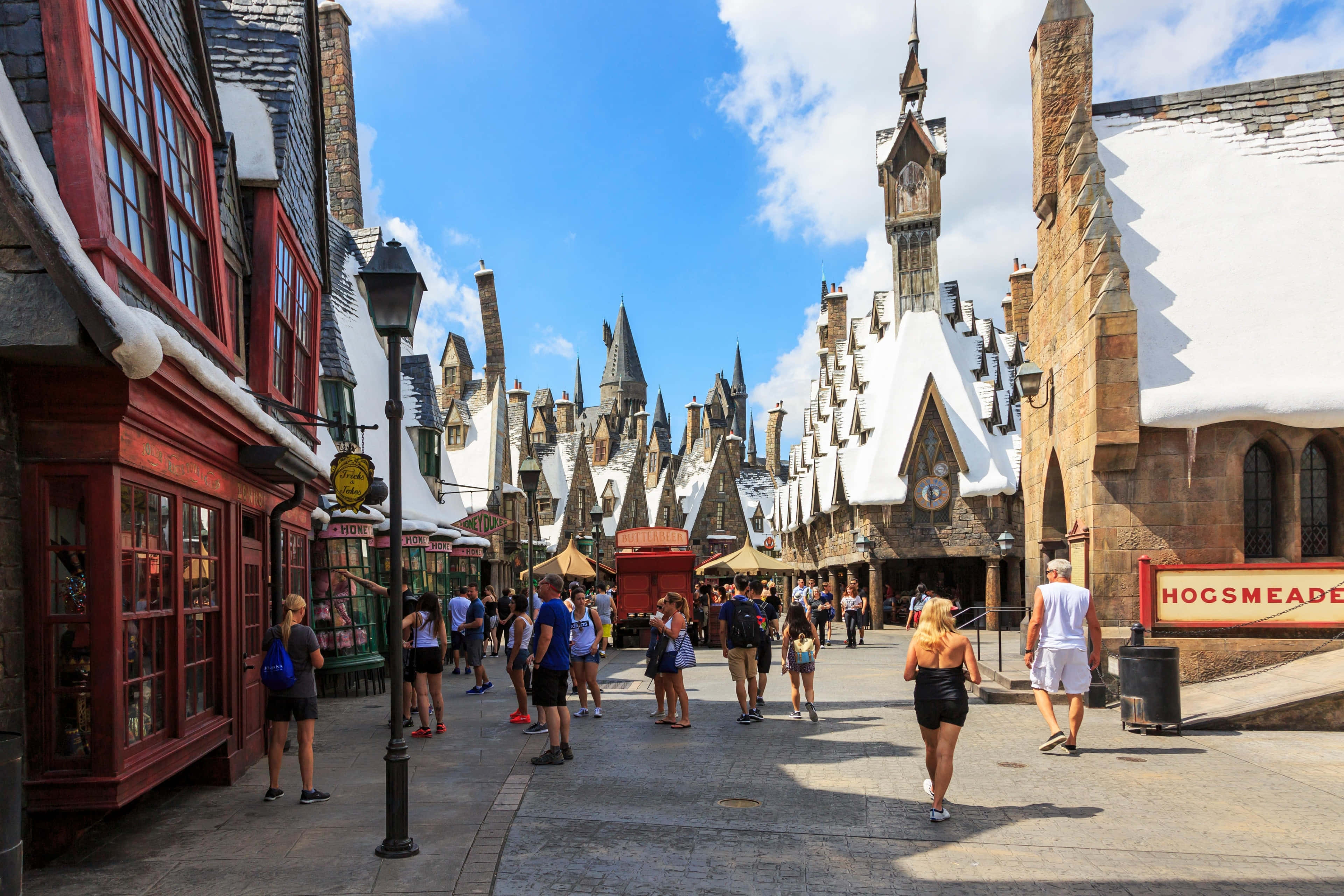 Wizarding World 桌布 壁紙 — 白雪覆蓋的中世紀風格建築排列在一條繁忙的鵝卵石街道上，上方是明亮的藍天和散落的雲朵。