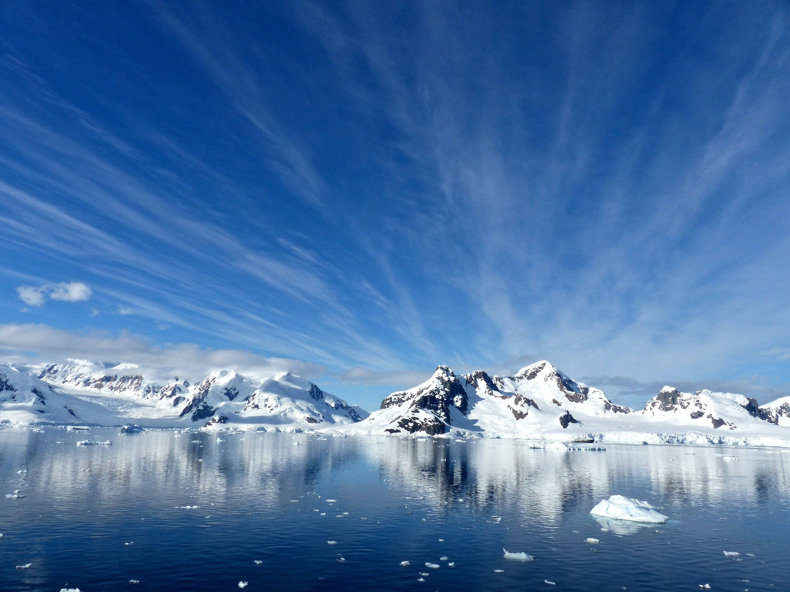 Antarctica 桌布 壁紙 — 白雪覆蓋的南極山脈倒映在平靜的藍色海水中，天空中有縷縷薄雲。