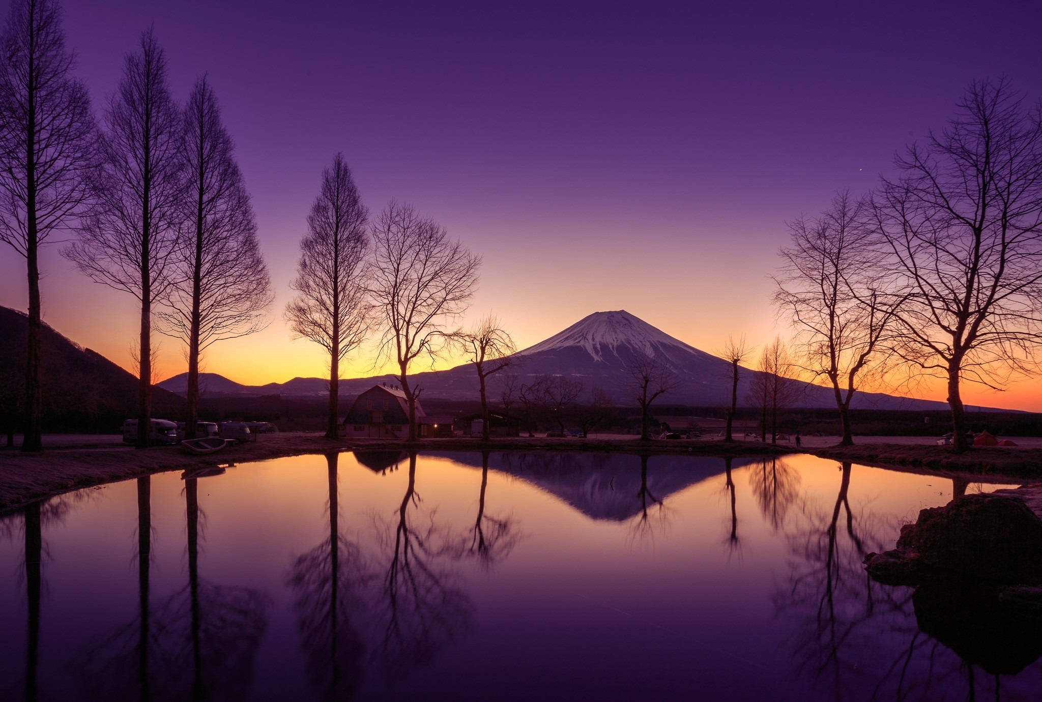 富士山 桌布 壁紙 — 白雪覆蓋的富士山倒映在寧靜的池塘中，池塘周圍是光禿禿的樹木，上方是充滿活力的紫色和橙色暮色天空。
