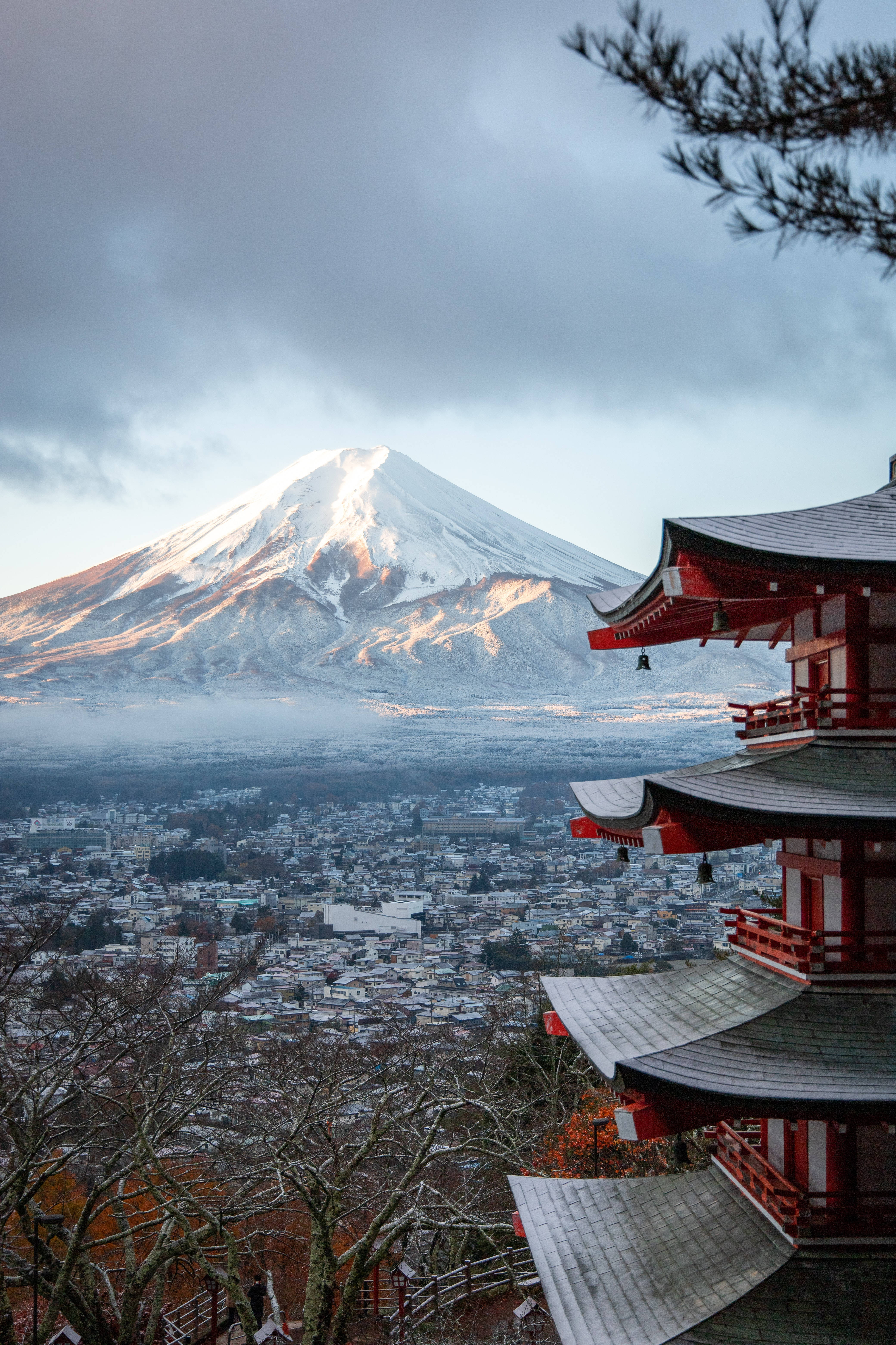 日本 桌布 壁紙 — 白雪覆蓋的富士山在傳統紅色寺廟背後雄偉地升起，寺廟的彎曲屋頂俯瞰著霧氣籠罩的小鎮。