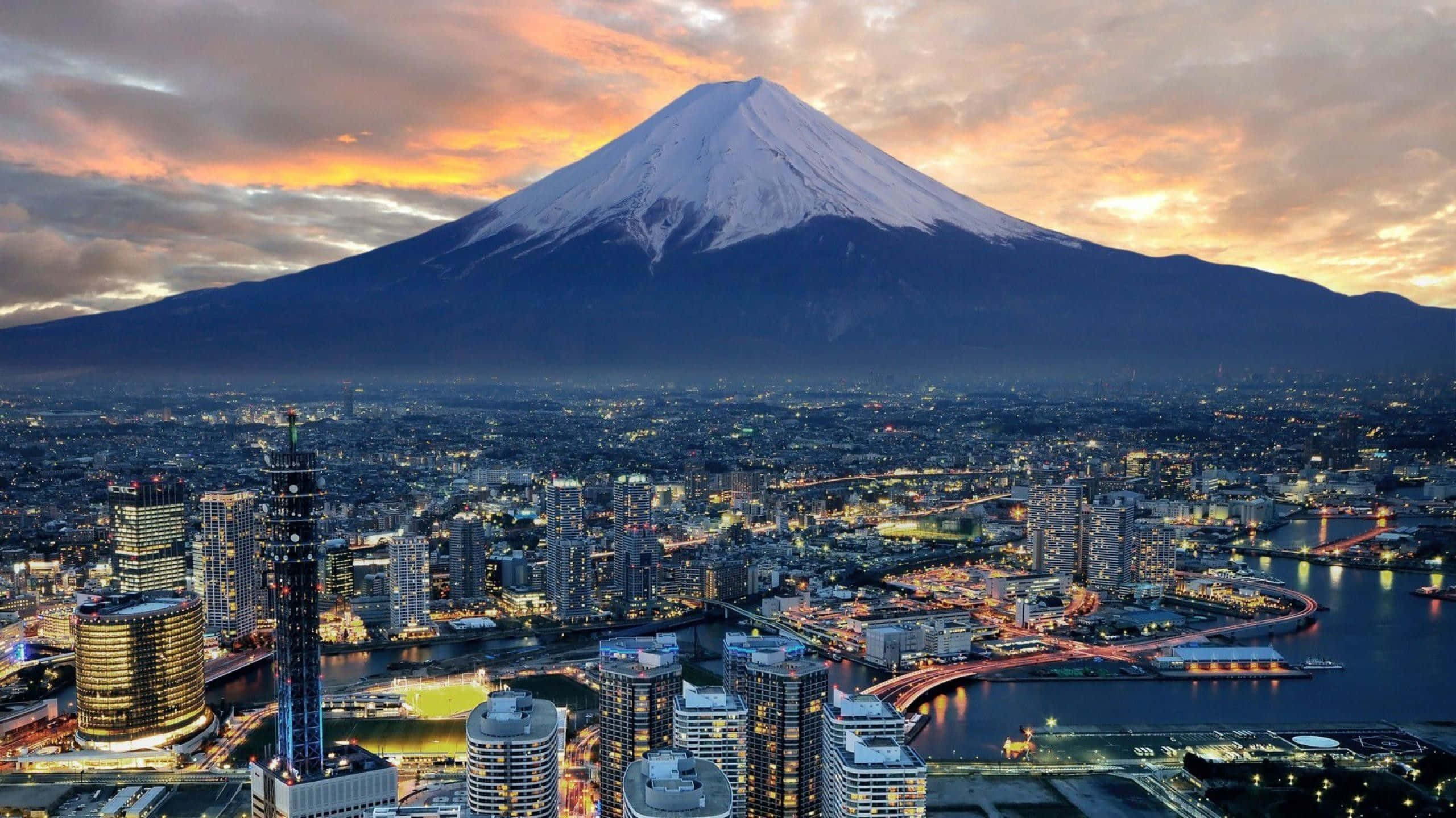 日本 桌布 壁紙 — 白雪覆蓋的富士山在日落時分聳立在發光的城市景觀之上，下方有溫暖的橙色雲彩和閃爍的燈光。