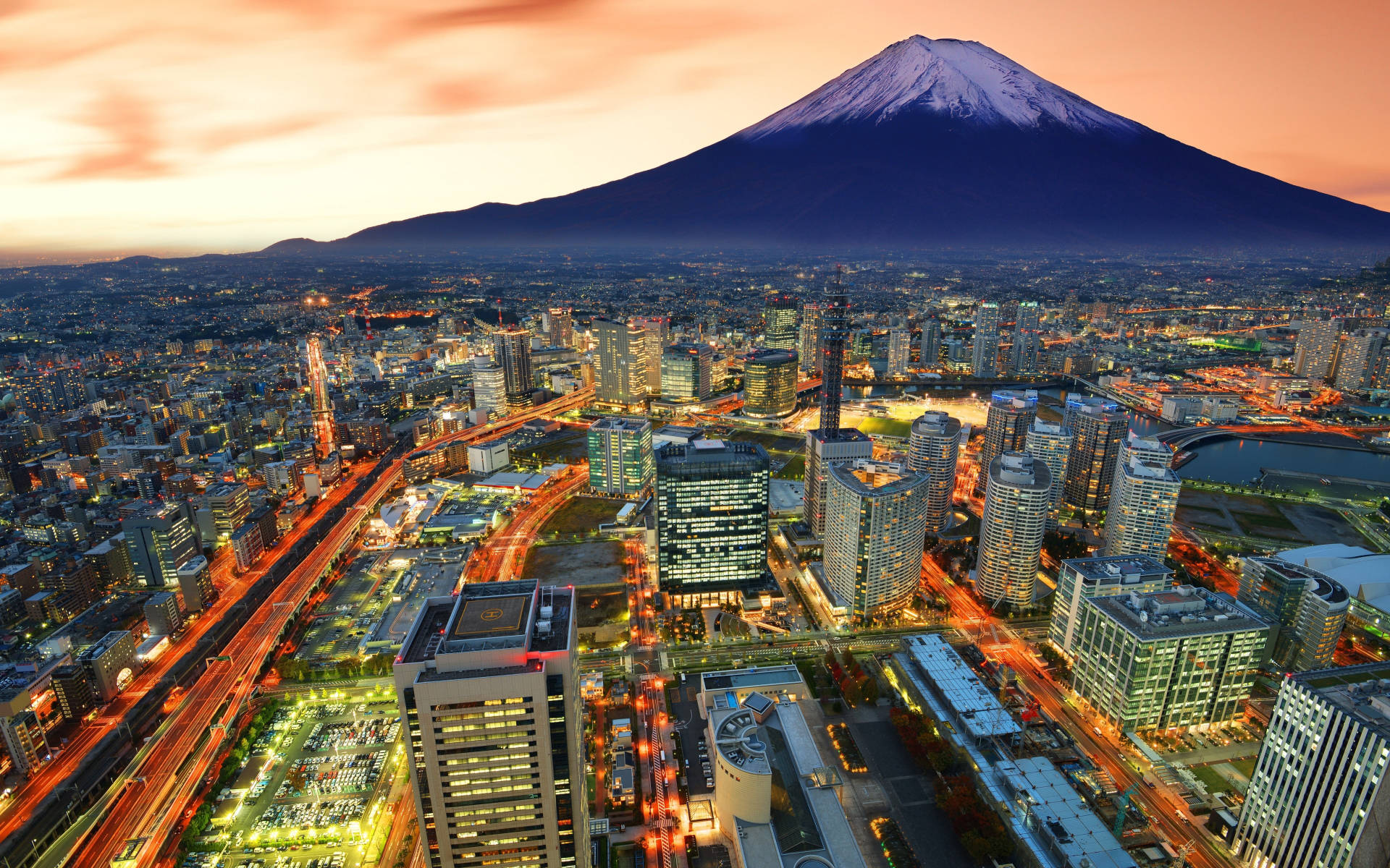 Yokohama 桌布 壁紙 — 白雪覆蓋的富士山在日落時分聳立於廣闊的城市景觀之上，伴隨著閃爍的燈光和流動的車流。