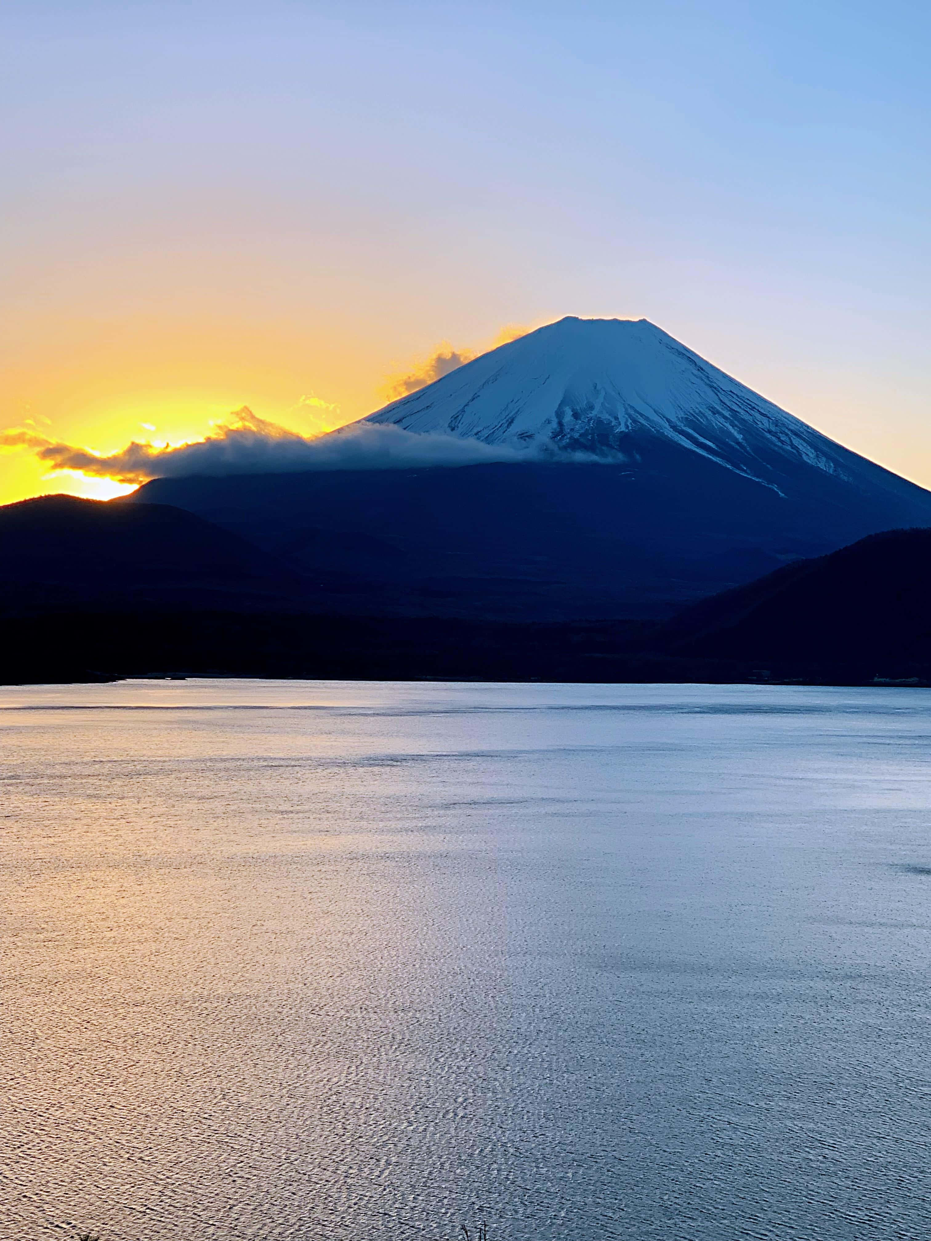 Mt Fuji 桌布 壁紙 — 白雪覆蓋的富士山在日落時分雄偉地聳立，山頂在漸變的天空下閃耀，倒映在下方平靜的水面上。