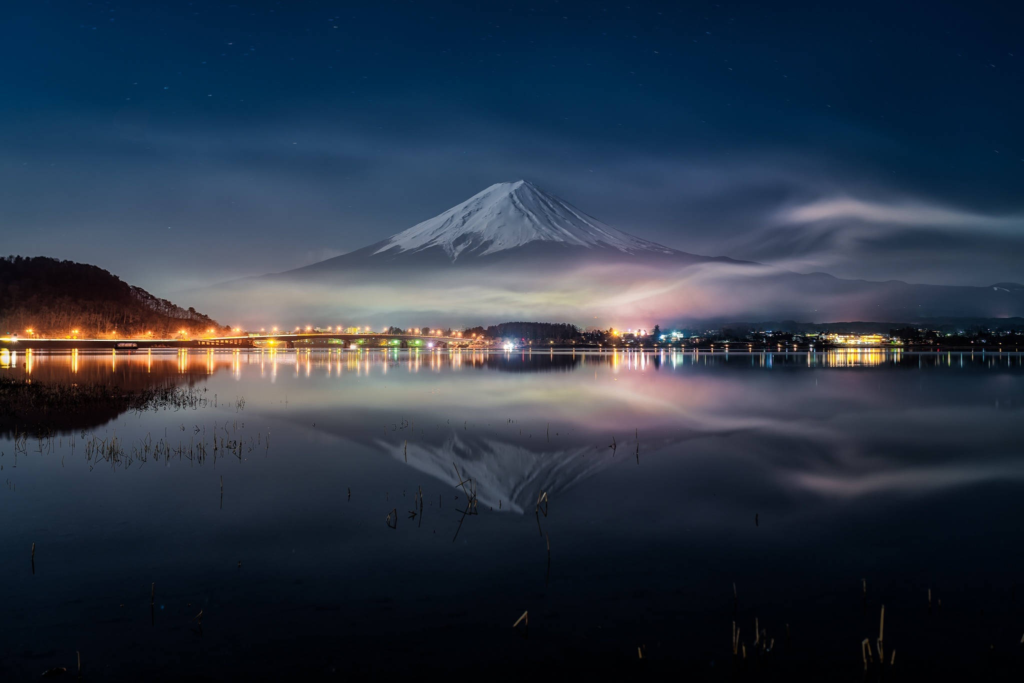 富士山 桌布 壁紙 — 白雪覆蓋的富士山在星光閃爍的湖面上平靜地倒映，城市燈光在深藍色夜空下在水面上閃爍。