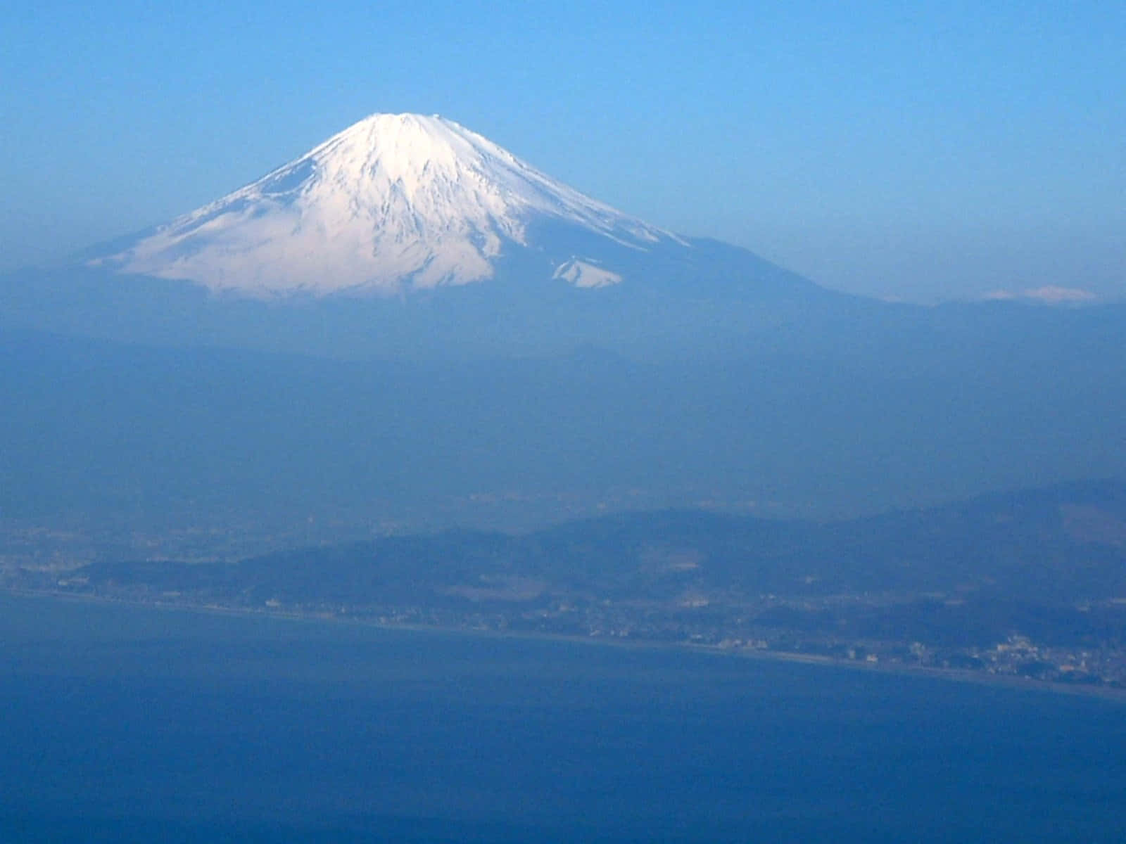 Mt Fuji 桌布 壁紙 — 白雪覆蓋的富士山在晴朗的藍天下，雄偉地聳立在朦朧的沿海平原之上。