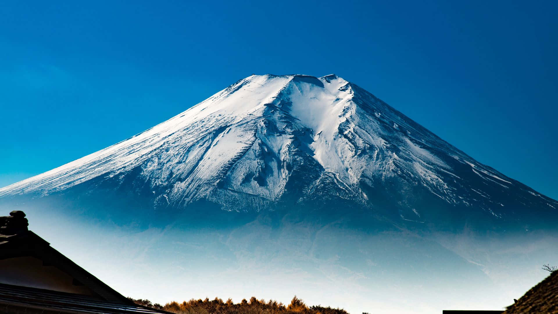 山脈 桌布 壁紙 — 白雪覆蓋的富士山在清澈的藍天下雄偉地聳立，前景可見傳統屋頂。