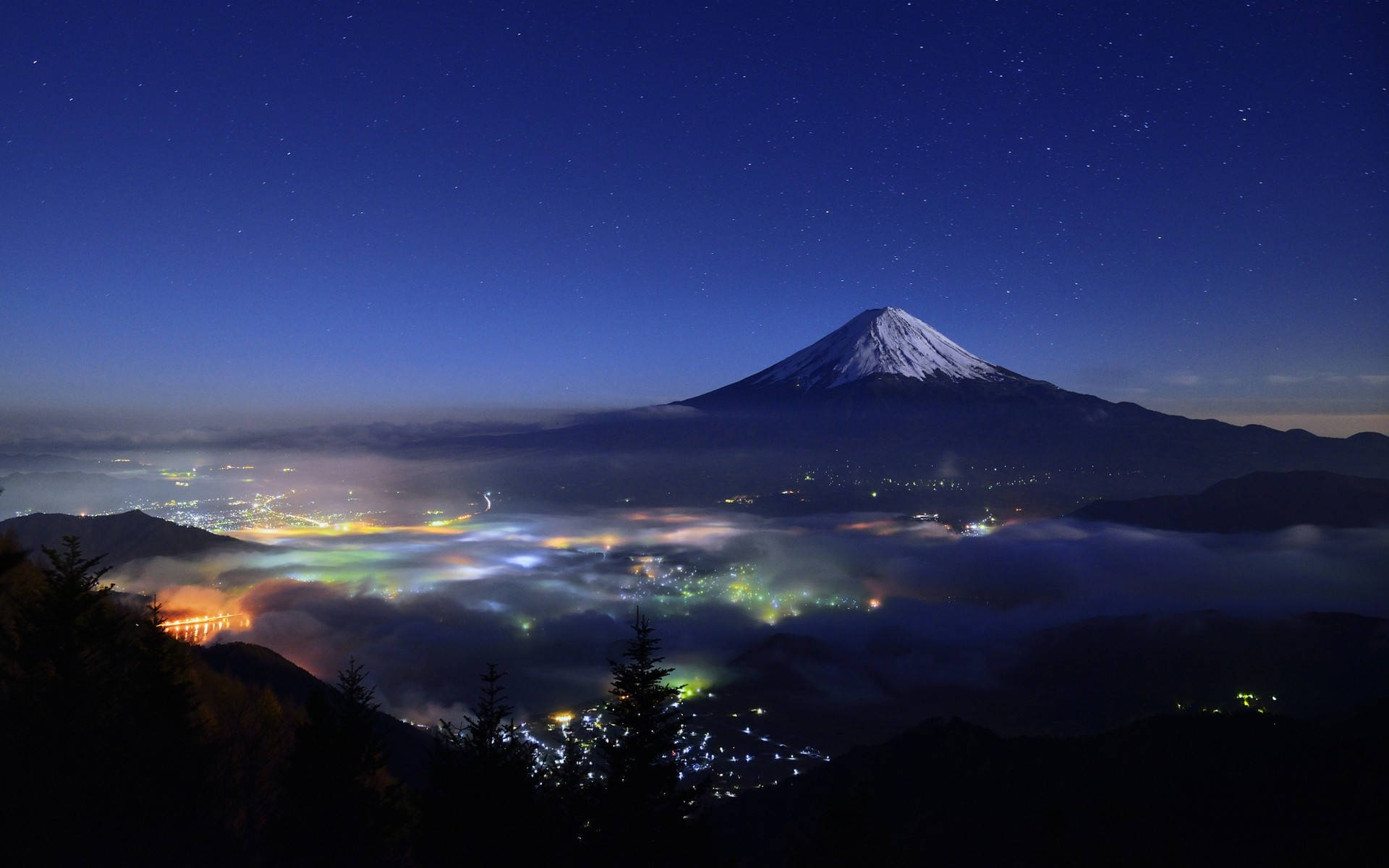 富士山 桌布 壁紙 — 白雪覆蓋的富士山在繁星點點的夜空下，聳立於霧氣籠罩的山谷中閃爍的城市燈光之上，氣勢磅礴。