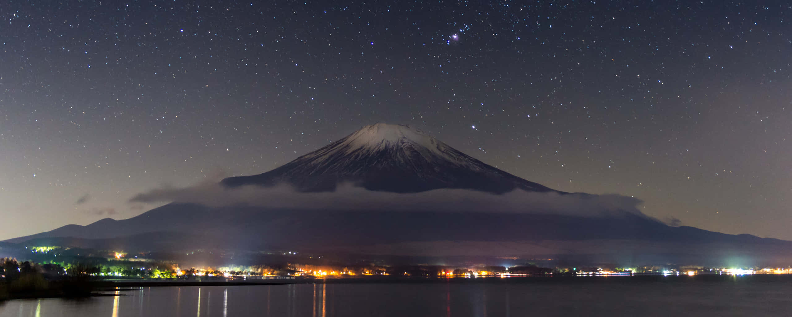 高清 桌布 壁紙 — 白雪覆蓋的富士山在繁星點點的夜空下雄偉聳立，城市燈光倒映在下方平靜的湖面上。