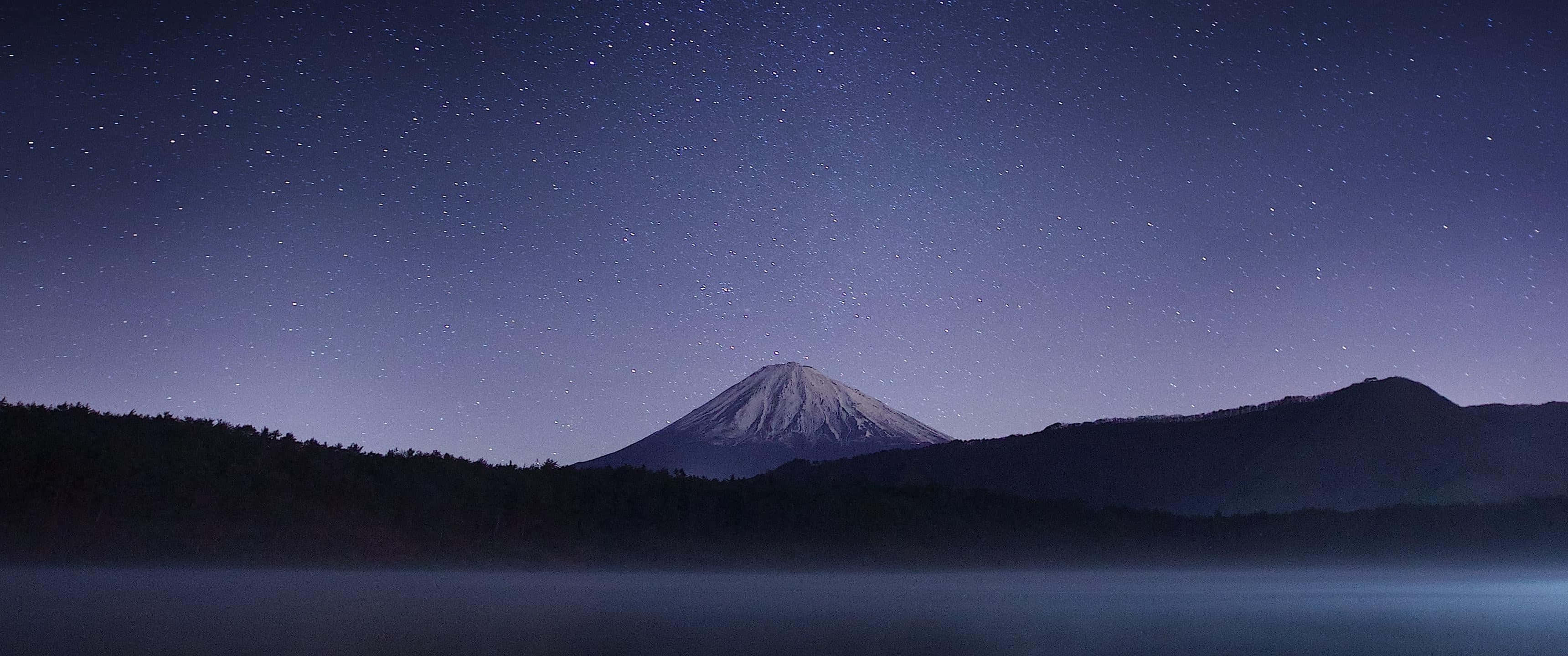 Ultra Wide 桌布 壁紙 — 白雪覆蓋的富士山在繁星點點的夜空下，寧靜地聳立在霧氣瀰漫的湖面上。