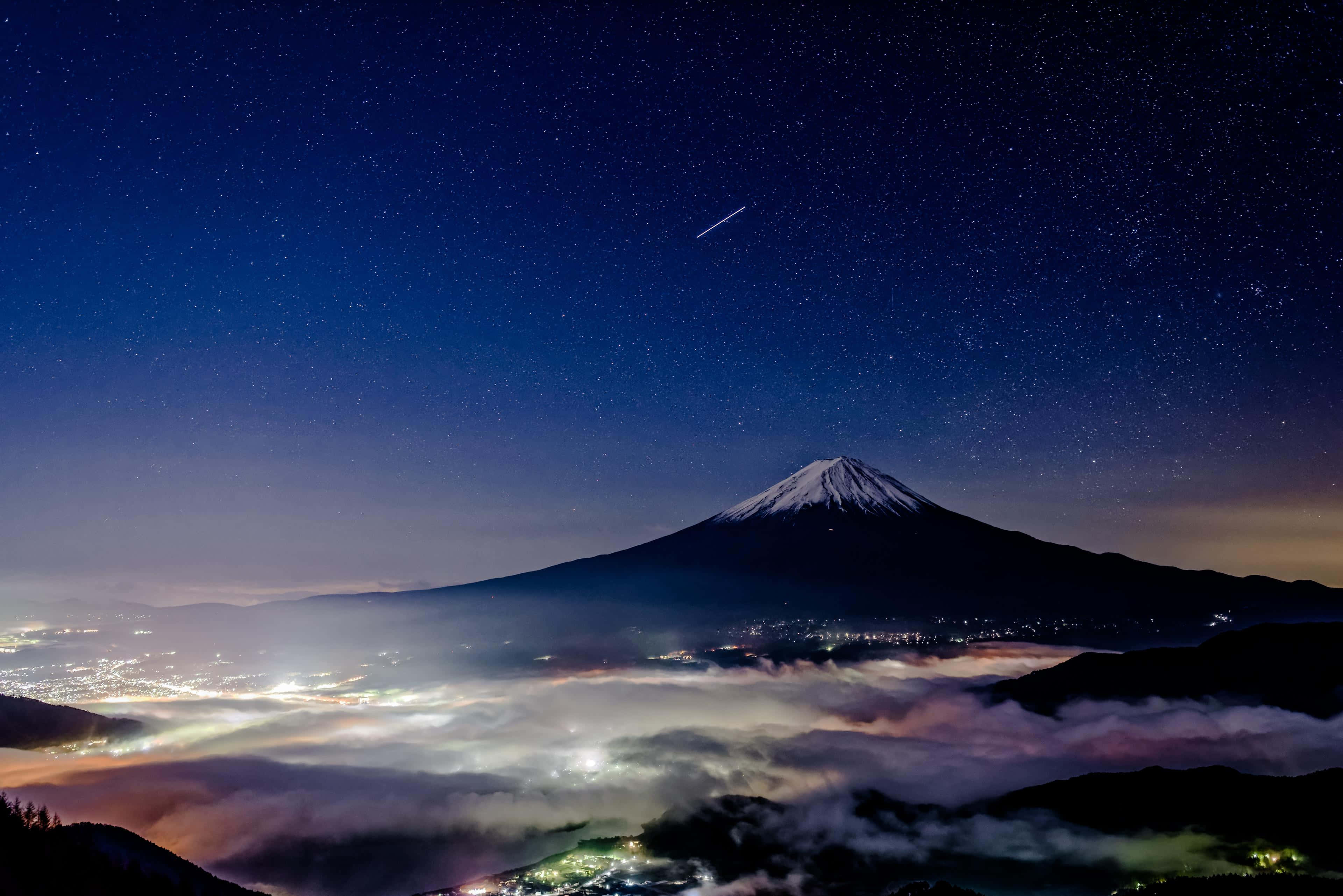 日本 桌布 壁紙 — 白雪覆蓋的富士山在繁星點點的夜空下巍然聳立，一顆流星劃過發光的雲海和遠處的城市燈光之上。